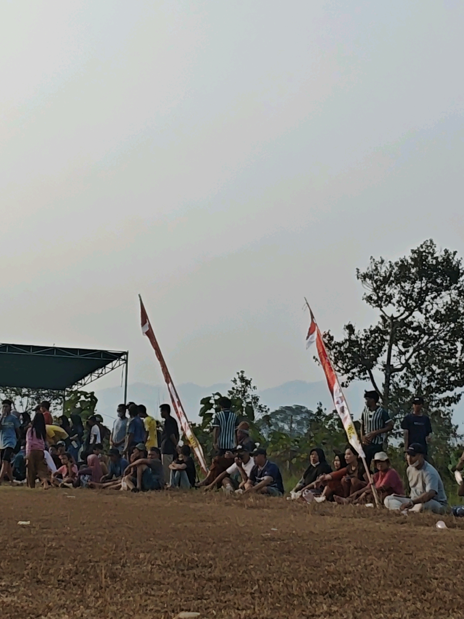 The image depicts an outdoor gathering of people, likely spectators, in the city of Karang Sari, Indonesia. The scene is set on a dry, brown, grassy field, with a backdrop of trees and distant mountains under a hazy, pale sky. In the foreground, a group of individuals are seated or standing, some engaged in conversation. Two flags, one predominantly red and white, are prominently displayed, held by individuals within the crowd. A temporary stage or shelter with a green roof is visible on the left side of the image. The overall impression is of a community event or sporting activity taking place outdoors.