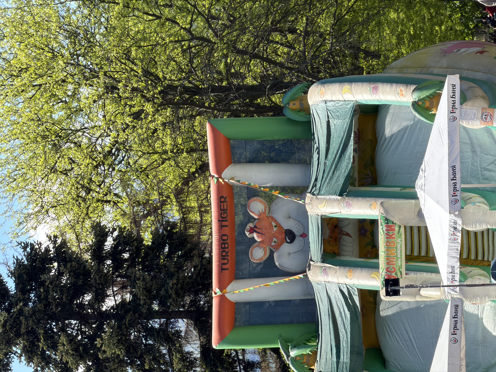 Here is a detailed description of the image:
**Scene:** The image depicts an outdoor inflatable bouncy castle in Sofia, Bulgaria. The bouncy castle has a "Turbo Tiger" motif and advertising for "Artski Center: Usmivki" (smiles) and "Gorna Banya" on its walls. Trees and blue sky are visible in the background.