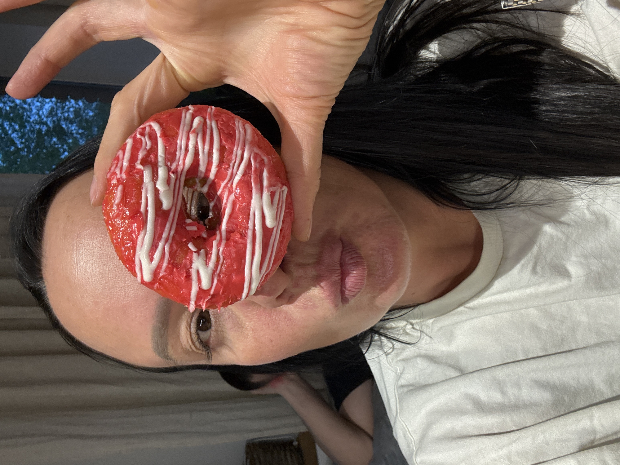 A woman is holding a red glazed donut with white drizzle in front of her face, partially obscuring her eye. She has dark hair and is wearing a white t-shirt. The background is out of focus and appears to be an interior setting with a window. There is no indication of the location being Chaikovsky, Russia.
