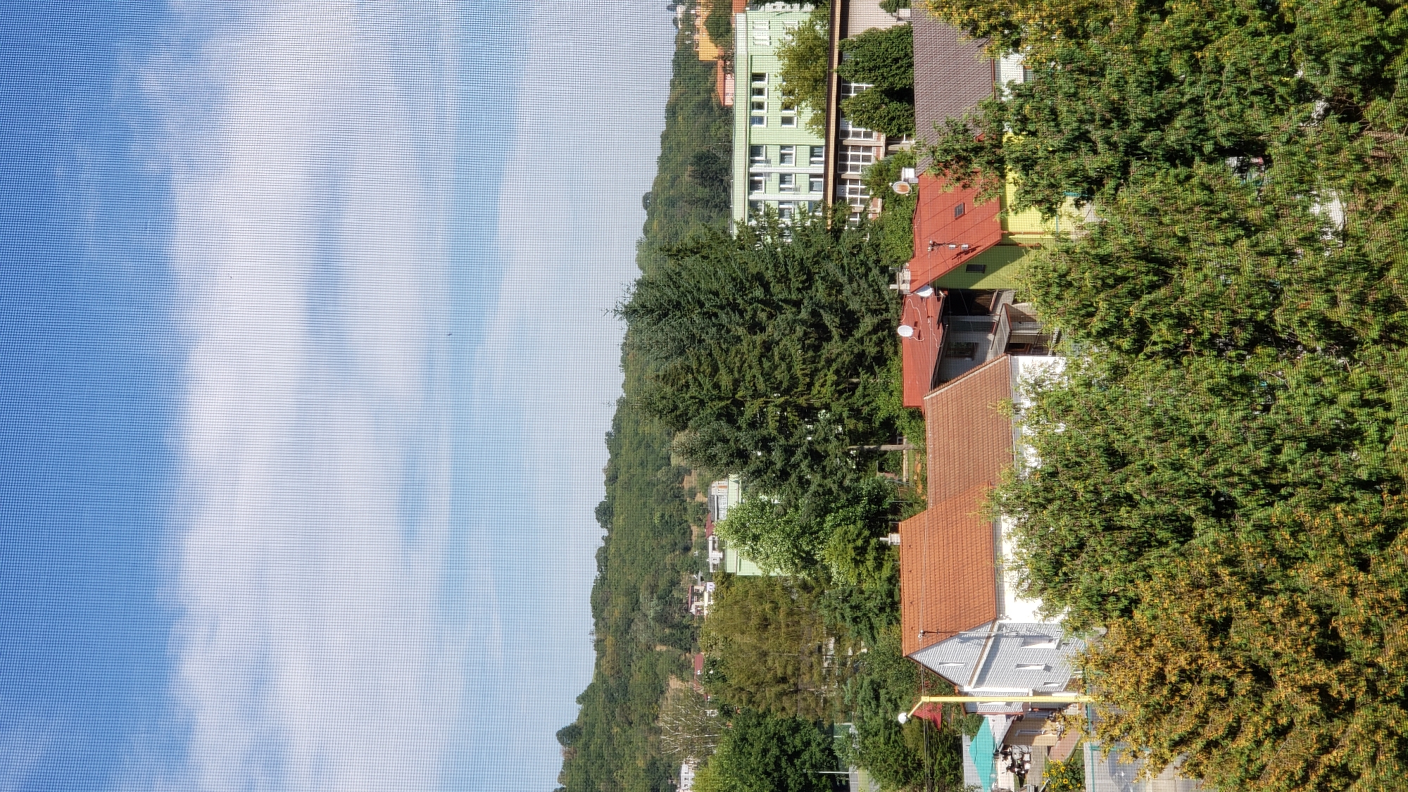The image depicts a residential area in Prešov, Slovakia, viewed from an elevated perspective, possibly through a window screen which creates a grid pattern over the scene. In the foreground, several houses with red tiled roofs are visible, partially obscured by lush green trees. One house to the left is white with a prominent gable. Adjacent to it, another house has a green facade and a red roof. Further back, a large, multi-story green building with numerous windows is prominent. Behind these buildings, a gently sloping, tree-covered hill forms the backdrop. The sky above is blue with scattered white clouds. The scene is set during daylight hours, suggesting an outdoor, daytime view of a neighborhood with a mix of residential buildings and abundant greenery.