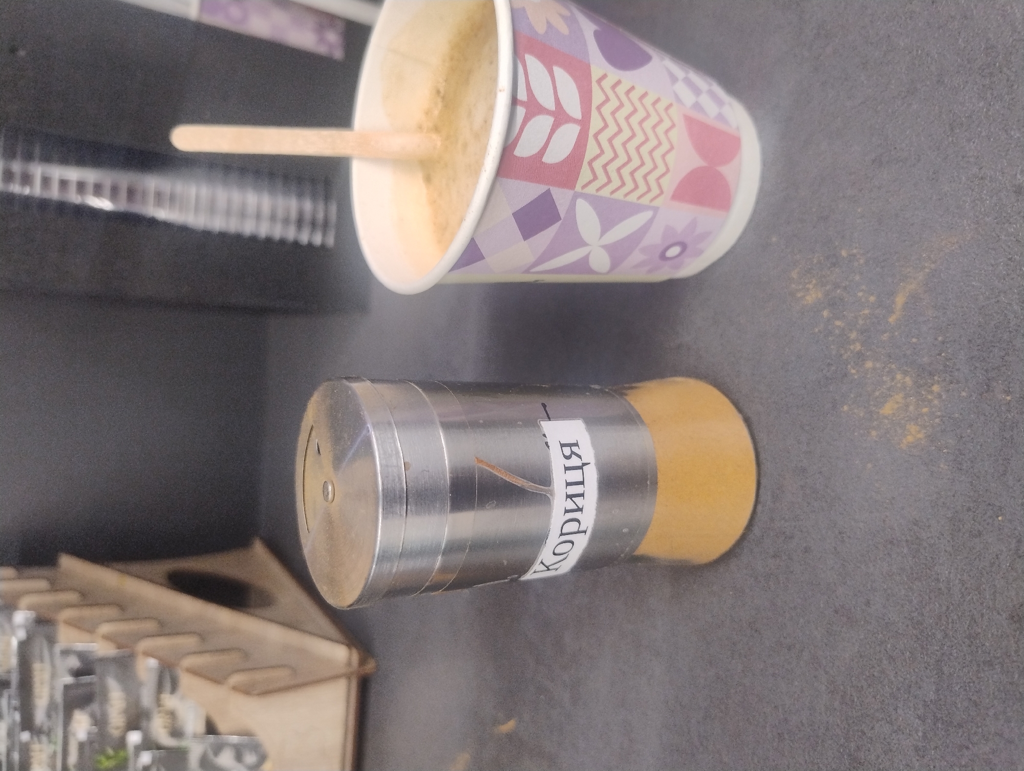 A close-up, slightly tilted overhead shot captures a table with a stainless steel spice dispenser labeled "Кориця" (cinnamon in Cyrillic) and a paper cup of coffee with a wooden stirrer. The spice dispenser is cylindrical with a metallic lid and a clear glass bottom filled with a yellow powder, likely cinnamon. The paper cup is decorated with a geometric pattern of pink, purple, and white shapes and contains a frothy, light brown beverage. A small pile of yellow powder is visible on the dark gray surface in front of the cup. In the blurred background, a wooden display rack holding several small packets is visible. The lighting is subdued, creating soft shadows.