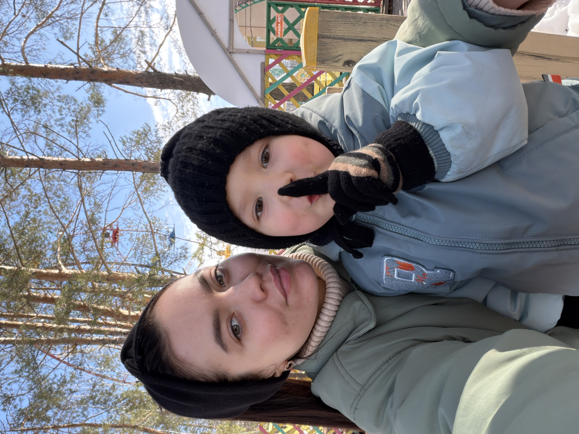A selfie-style portrait captures an adult female and a young child outdoors on a sunny winter day in Chaikovsky Park Kultury i Otdykha, Chaikovsky, Russia. The adult female, positioned on the left, wears a light olive-green puffer jacket over a beige turtleneck sweater and a black headband. She has dark hair, brown eyes, and is looking directly at the camera with a slight smile. The child, located on the right and slightly in front, wears a black knitted beanie hat, a light blue puffer jacket with a grey patch featuring an orange 'P' on the left sleeve, and black gloves with horizontal beige stripes. The child holds their right index finger vertically to their lips in a "shhh" gesture, also looking directly at the camera. The background features tall pine trees against a clear blue sky with sparse white clouds. Visible elements of an amusement park include red and blue gondolas from a Ferris wheel, a colorful wooden lattice fence (green, yellow, pink, white), and a white building with a snow-covered roof. A green sign with white text, partially visible, reads "ВХОДА НЕТ" (NO ENTRY). The ground appears to be snow-covered, consistent with winter conditions.