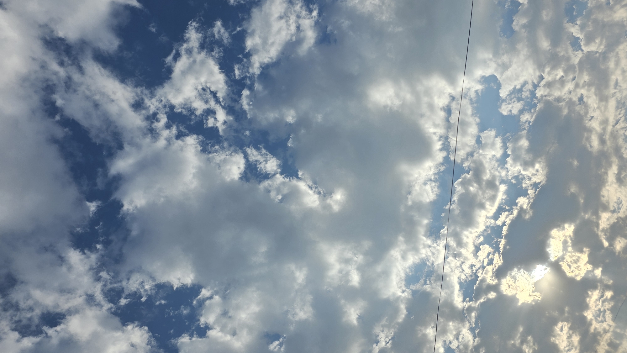 An outdoor, vertical, eye-level shot captures a vast expanse of sky filled with clouds, with the sun partially obscured by cloud cover. The clouds are a mix of bright white and darker gray, creating a dynamic contrast against the deep blue of the sky. The sun's rays are breaking through the clouds, casting a warm, ethereal glow. A thin, dark wire stretches horizontally across the middle section of the image, suggesting the presence of power lines or telephone lines. The scene evokes a sense of openness and natural beauty. The image is set in the city of Chuy, Kyrgyzstan.