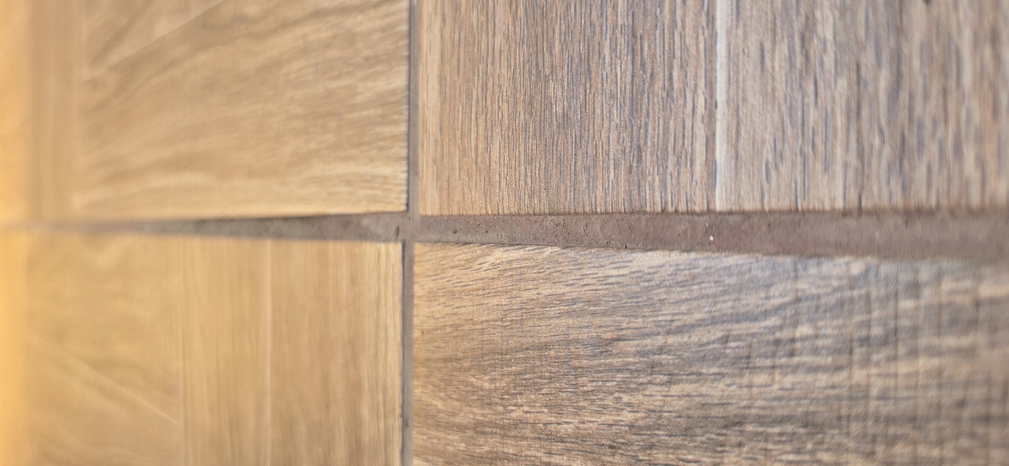The image shows a close-up, eye-level view of a floor with a wood-like tile pattern. The floor is composed of four square tiles meeting at a central grout line. The lighting appears to be warm, possibly from an overhead source. The tiles exhibit variations in color and wood grain texture, with some appearing lighter and smoother, while others are darker and more pronounced in grain. The grout line is a reddish-brown color and appears to be slightly uneven. There are no people, animals, or other objects visible in the frame, and the scene is static. The location in Urupskiy, Russia, is not discernable from the image itself.