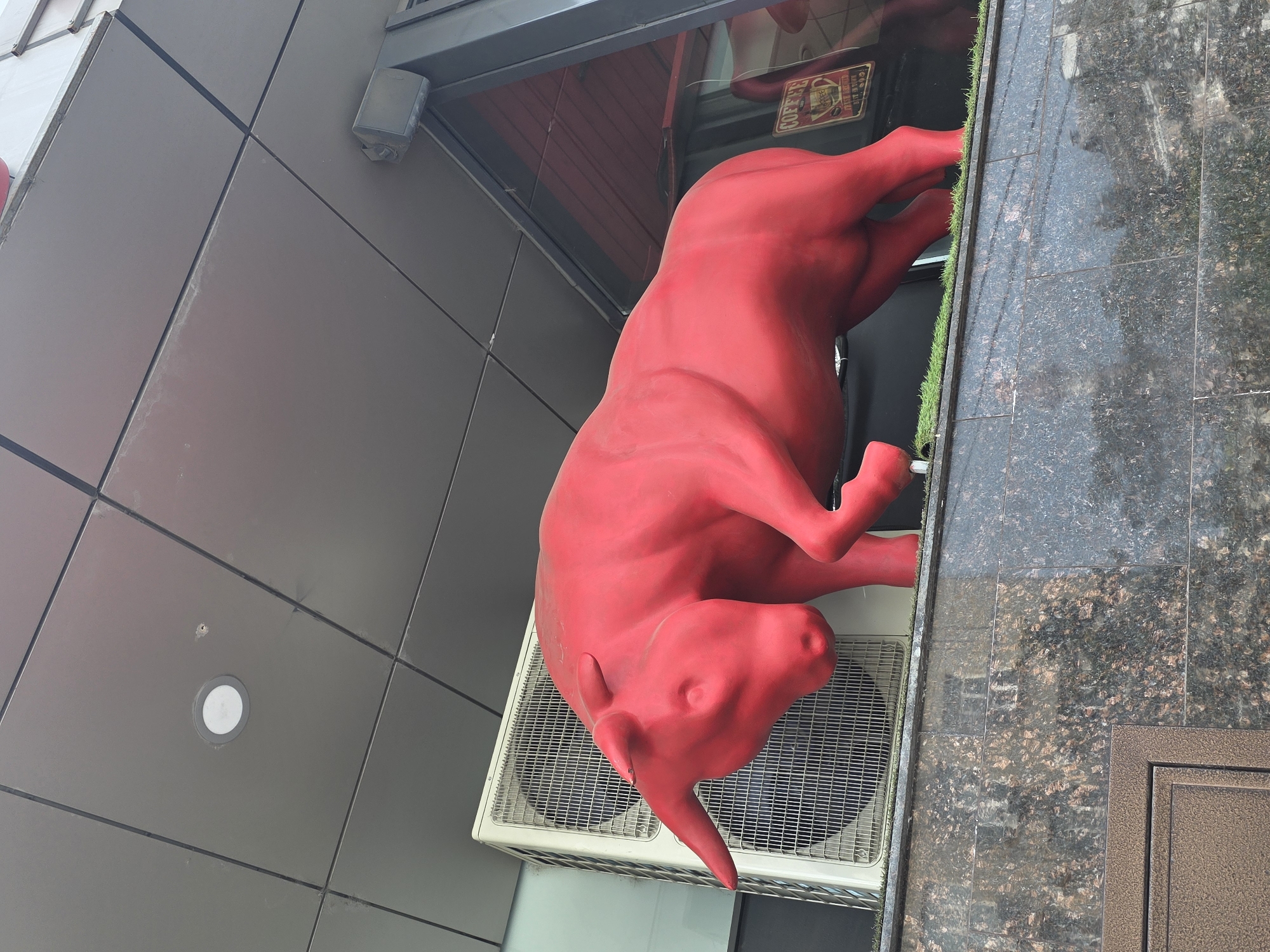 A full-body statue of a bright red bull is mounted on top of what appears to be an air conditioning unit attached to the side of a modern building with grey paneling. The bull is in a reclining position, with its front legs slightly tucked and hind legs extended. Part of a storefront can be seen above the bull, including a sign with the word "COFFEE" visible in reverse. The building is set against a cloudy sky. Next to the air conditioning unit are tiled surfaces and brown metal trim.
This appears to be in Krasnodar, Russia, based on information related to similar statues and locations.