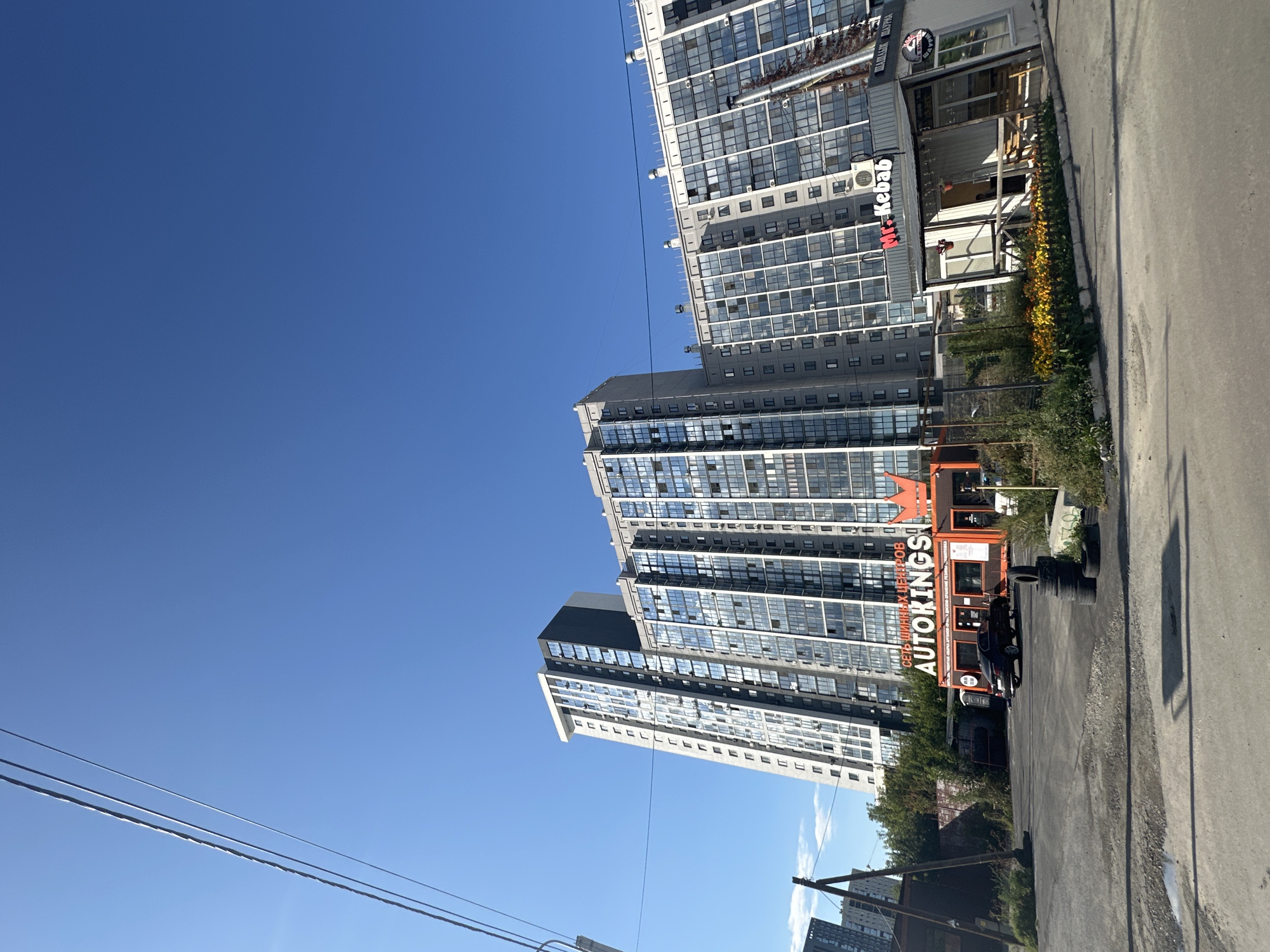 In a wide-angle shot, a modern, multi-story residential building with numerous windows dominates the background, its facade reflecting the clear blue sky. In the foreground, a small food kiosk with a sign reading "Mr. Kebab" is visible, along with a red structure labeled "AUTOKINGS" which appears to be a car service center. A black car is parked in front of the "AUTOKINGS" building, and several tires are stacked nearby. The environment suggests an urban setting with overhead power lines crisscrossing the sky.