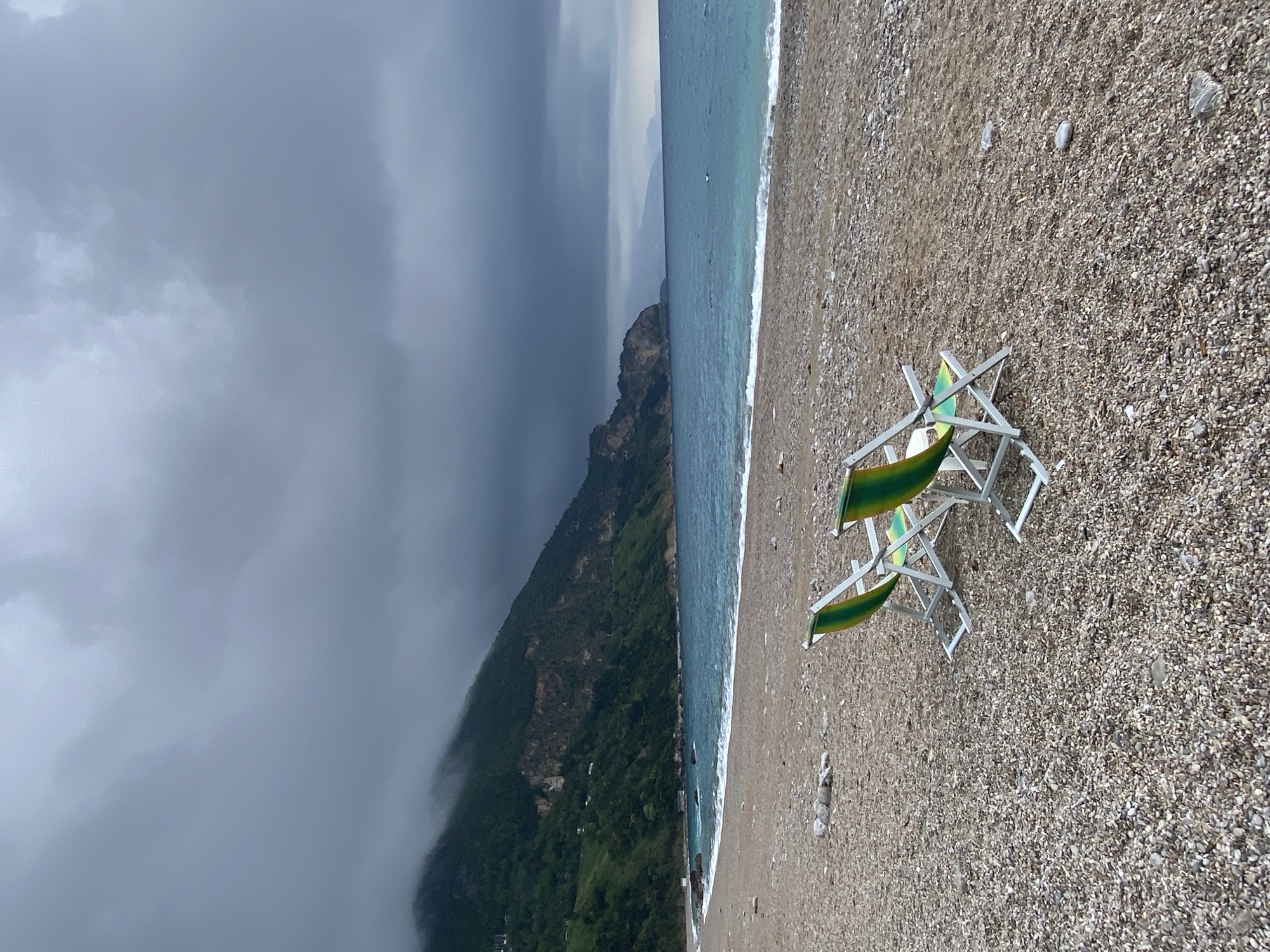The image shows a beach scene in Prijevor, Montenegro, with two green and white beach chairs on a pebble beach. In the background, a mountain slope is partially obscured by low-lying clouds or fog. The sea stretches along the coastline, meeting the beach with small waves. The sky is overcast with dark, heavy clouds.