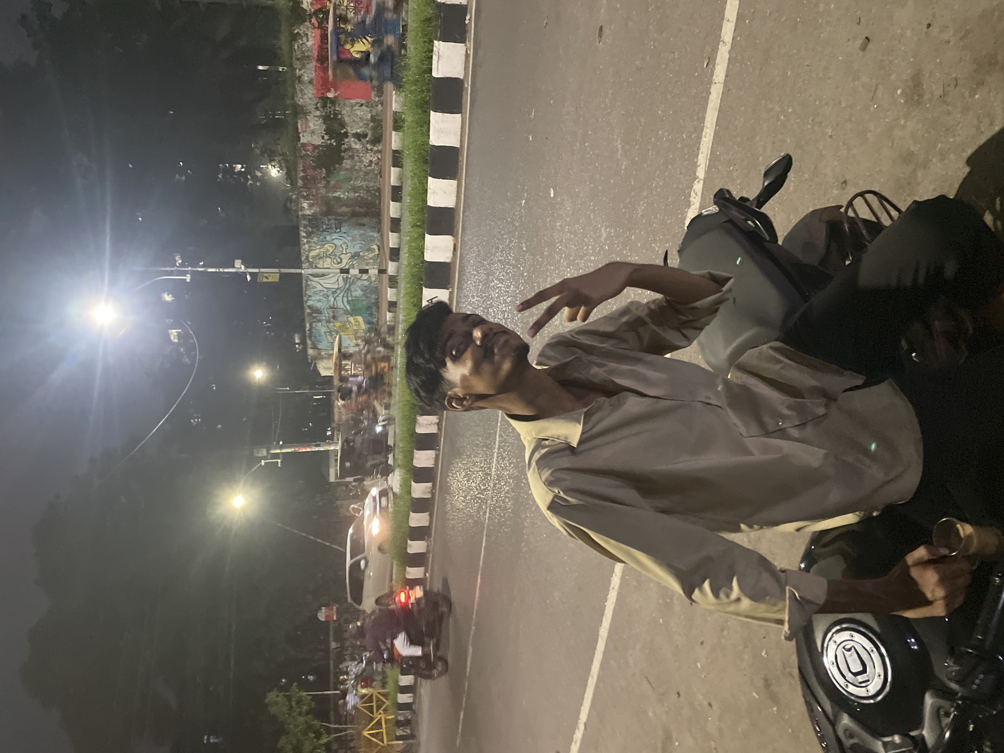 A person on a motorcycle is photographed at night in Dhaka, Bangladesh. The individual, wearing a light-colored button-down shirt, is partially visible and making a peace sign. The motorcycle is black and positioned on the right side of the frame.
The backdrop features a street scene with moving traffic. A wall with graffiti is visible, bordered by a black and white striped curb. Streetlights illuminate the area. In the distance, other vehicles and people can be seen, contributing to a sense of urban activity.