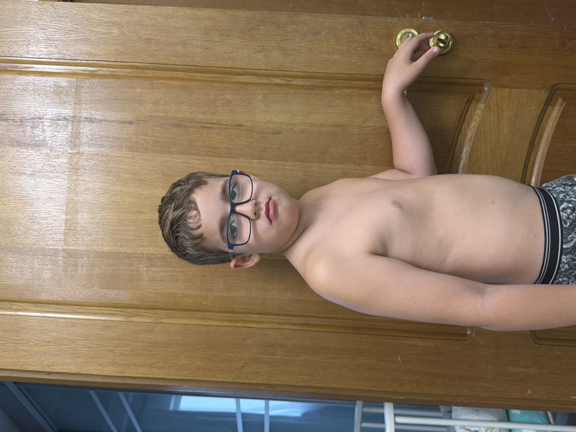 A shirtless boy with short brown hair and blue-framed glasses is lying on a wooden door, grasping the doorknob with his right hand. He is wearing patterned boxer shorts with a black and white design. The door has a horizontal grain pattern and a decorative arched panel on the right side. Part of a window with white frames is visible at the bottom of the frame. It is not possible to determine if this image was taken in Sochi, Russia.