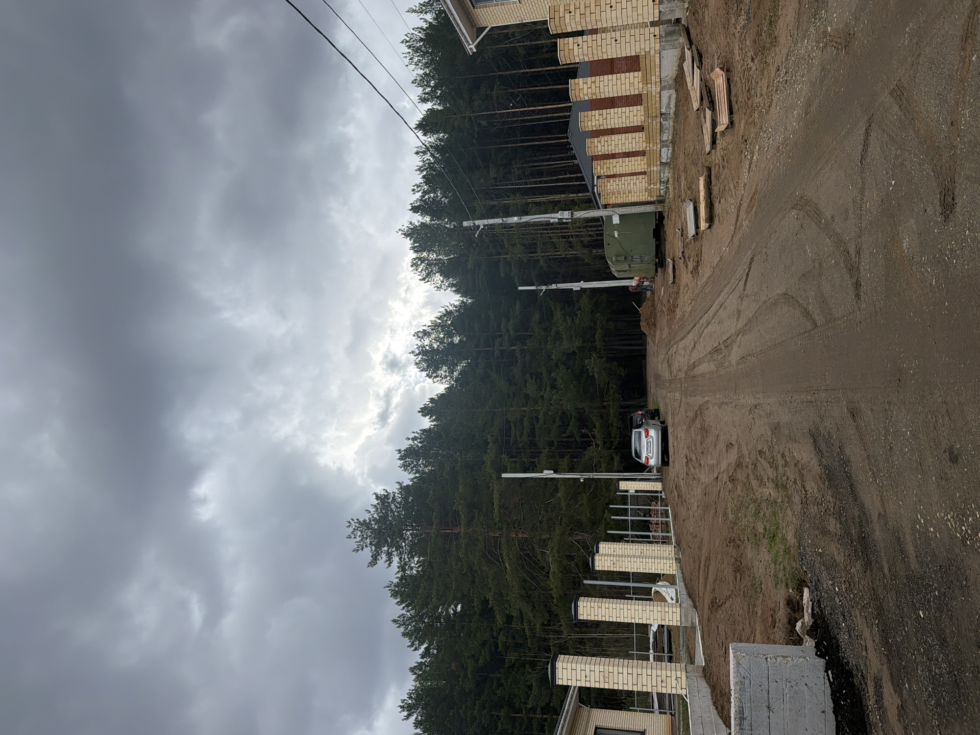 The image captures an outdoor scene under a heavily overcast, cloudy sky, suggesting diffused daylight. A wide, unpaved dirt road or path stretches from the foreground towards the background, curving slightly. The ground is rough and uneven, marked with visible tire tracks.
To the left of the road, a dense forest of tall, dark green pine trees forms a prominent backdrop. Overhead power lines run horizontally across the upper left portion of the image. Two tall, white poles, possibly utility or street light poles, stand between the road and the forest. A green utility vehicle, possibly a truck with a boom lift, is parked near the forest edge. A person wearing an orange hard hat is partially visible near this vehicle, indicating work in progress. Further down the path, a white car is parked facing away from the viewer.
To the right of the road, various construction elements are visible. These include multiple light-colored brick pillars, possibly part of a fence or gate, and sections of metal framing. A building, constructed from light-colored bricks with darker brick accents, is partially visible. Wooden planks are laid on the ground near these structures. A concrete block is visible in the immediate foreground on the right.
The scene depicts an area under development, blending natural forest elements with ongoing construction in what appears to be a semi-rural or suburban setting in Chaikovsky, Russia. No legible text is visible.