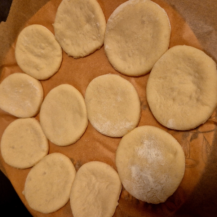 Eleven doughy circles, possibly cookies or biscuits, are arranged on a round baking sheet lined with parchment paper. The circles vary in size, some being larger than others. Their surfaces are pale yellow and slightly dusted with flour. The parchment paper is light brown. The baking sheet beneath is dark yellow.
