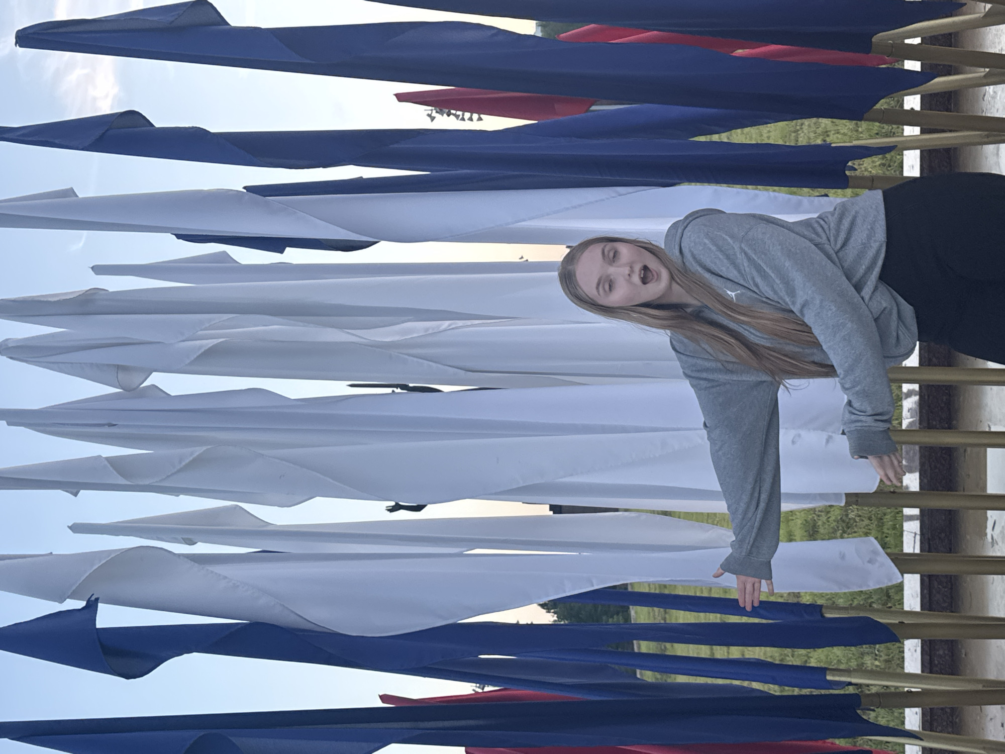 A young Caucasian girl with blonde hair is leaning against a row of flags. She is wearing a gray hooded sweatshirt and dark pants. Her mouth is open, and she appears to be making a surprised or playful expression. The flags are in alternating colors of blue, white, and red, and they are positioned in a staggered arrangement. The scene is outdoors, with a clear sky in the background. The time of day appears to be late afternoon or early evening, suggested by the warm, soft light. The location could be a park or an outdoor venue decorated with these patriotic or festive flags. There is no visible text in the image. The overall mood is lighthearted and energetic, captured by the girl's animated pose and expression.