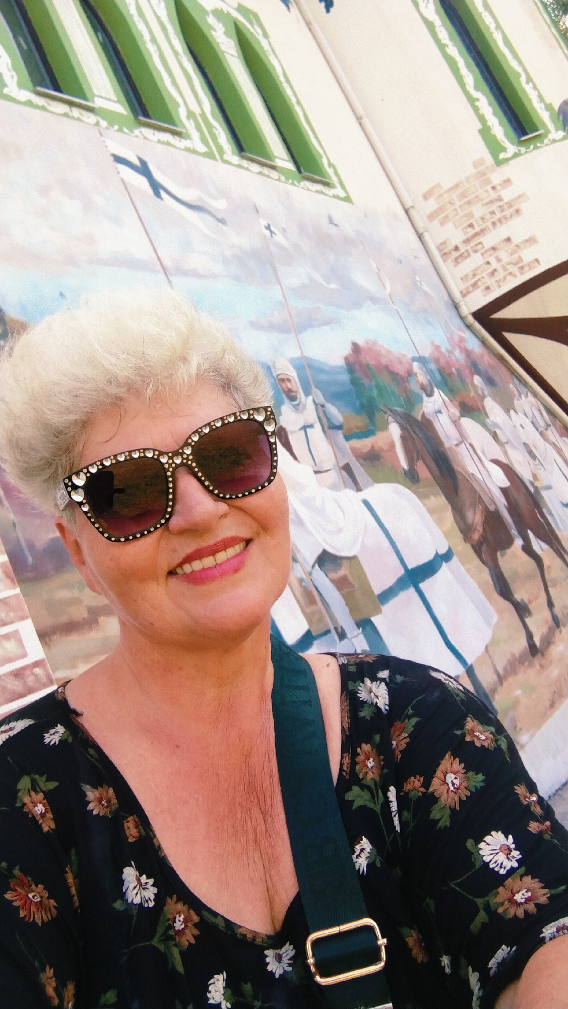 A selfie features a middle-aged woman with blonde hair, wearing oversized sunglasses adorned with rhinestones and a black floral-print dress. She is smiling and looking directly at the camera. Behind her, a colorful mural depicts a medieval scene with knights on horseback. The background also shows elements of a building with arched green windows. The image appears to have been taken outdoors in the city of Horishni Plavni, Ukraine, likely near a public mural or landmark.
