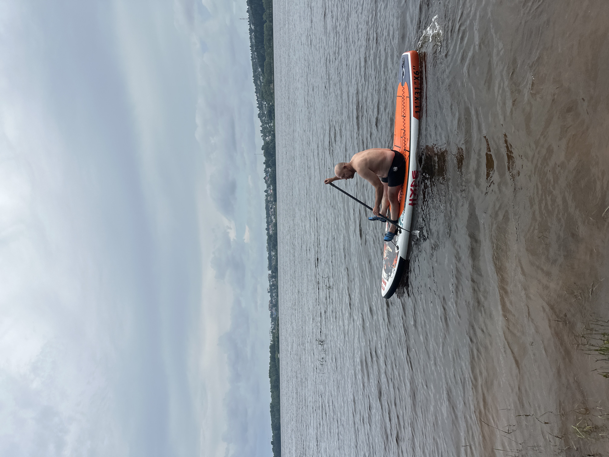 An overhead, medium shot captures a bald, shirtless man paddleboarding on a large body of water under an overcast sky. The man is seated on an orange and white paddleboard, holding a paddle. The water is rippled, and the shoreline with trees and buildings is visible in the distance. The scene appears to be the city of Noviy, Russia, based on the context provided.