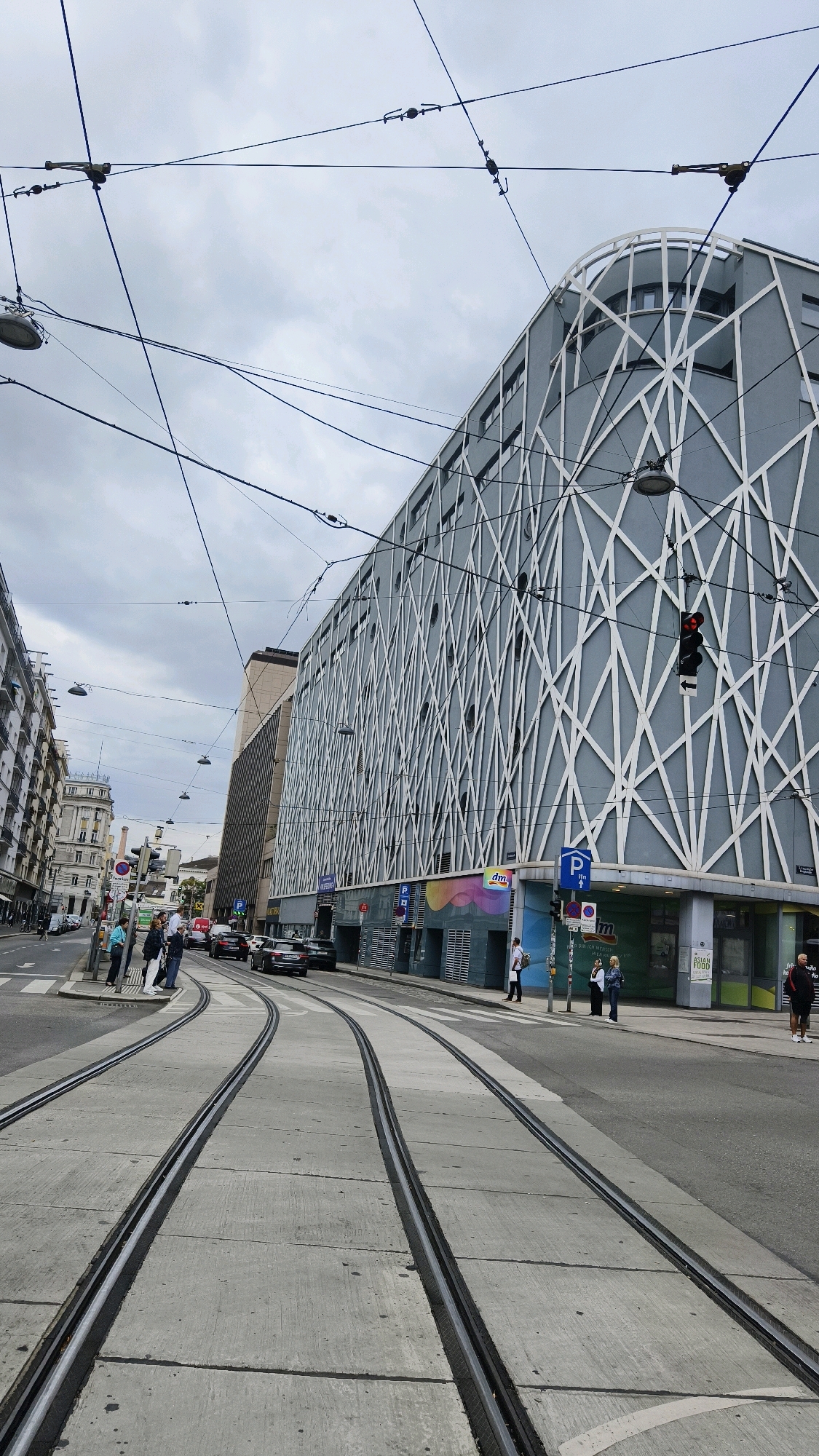 The image depicts a street scene in Vienna, Austria, with tram tracks prominently featured in the foreground. On the right side of the street is a modern, grey building with a white, lattice-like facade. Several signs are visible on this building, including a blue parking sign and a sign for "dm." To the left of the tram tracks, a row of older, multi-story buildings lines the street, with awnings and balconies. There are several people on the sidewalk and waiting near the tram tracks, and a few cars are visible on the street. Overhead, a complex network of tram wires crisscrosses the sky, along with streetlights and traffic signals. The sky is overcast.
