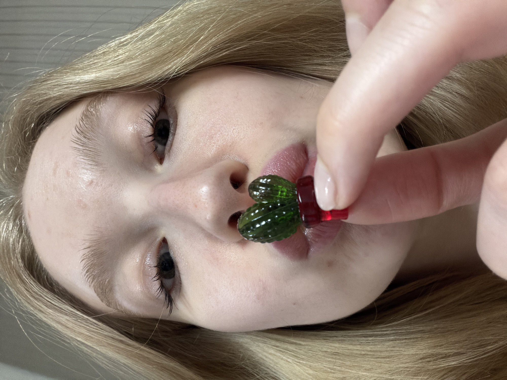 A young, blonde woman is holding a cactus-shaped gummy candy between her lips. Her face is fair and she has long eyelashes. The scene is indoors, the background is indistinct. There is no indication of the location being Yuzhny, Russia.