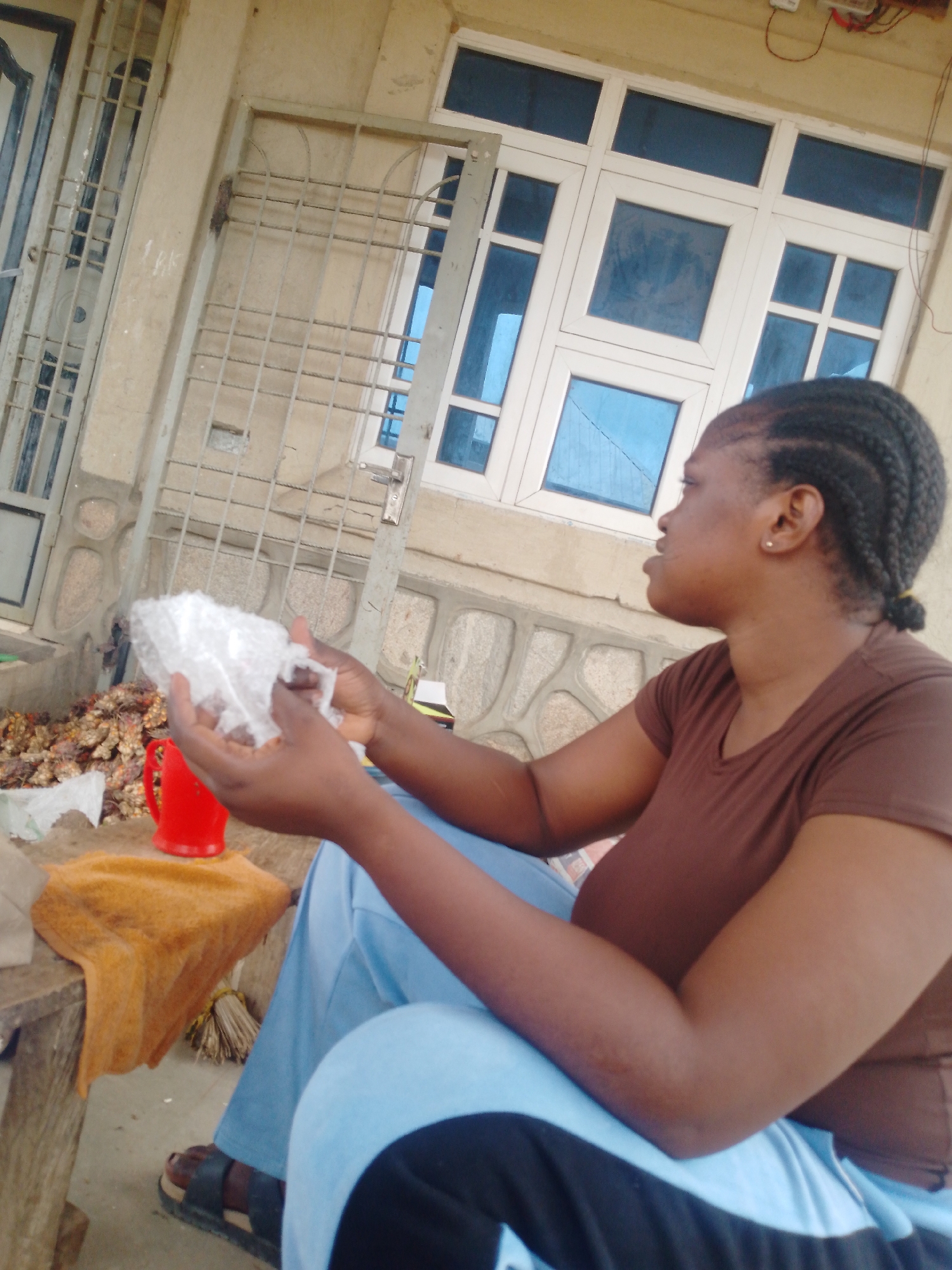 A medium-shot image shows a woman seated outdoors or in a semi-enclosed space, looking right in profile. She has dark skin, braided hair pulled back, and is wearing a plain brown t-shirt and light blue pants. Her hands are holding a clear, crinkled plastic bag or wrapper, which appears to contain a white or light-colored, lumpy substance.
To her left, on a low wooden stool or table, a bright red plastic cup is visible. Next to the cup is a pile of what looks like dried turmeric roots or similar spices, some of which are partially covered by an orange-brown cloth. A small, tied bundle of sticks, possibly a broom, rests beneath the cloth.
The background features a building facade with a large white window frame containing multiple panes, some of which reflect a blue sky or surroundings. A metal security gate or grill covers another section of the wall, which is light-colored and has textured stone-like elements at its base. In the upper right corner, electrical wires and what appears to be an electrical meter or box are mounted on the wall.
The lighting suggests it is daytime. The woman's expression is neutral, and there is no visible text. The overall setting implies a casual, possibly commercial or domestic, outdoor environment, perhaps in a warm climate.