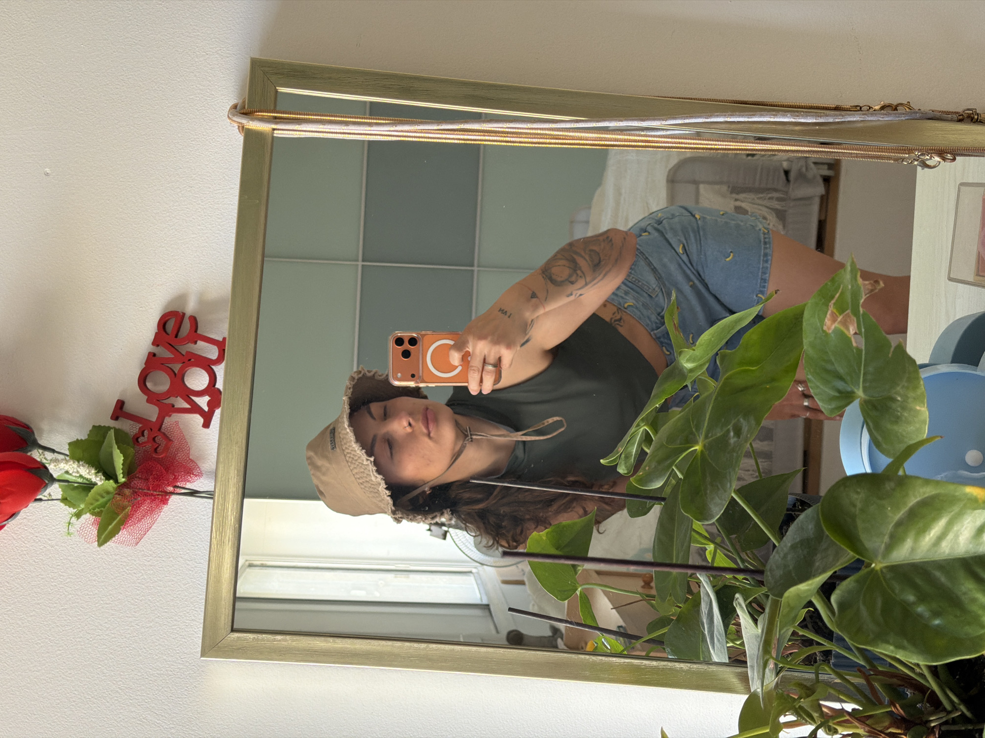A reflection in a gold-framed mirror shows a woman with medium-length curly brown hair wearing a tan bucket hat, a dark green t-shirt, and blue jean shorts with yellow banana patterns. She is holding an orange smartphone with a white ring on the back. Her left arm, visible in the mirror, has multiple tattoos. In the foreground, a green plant with large leaves partially obscures the view. To the left of the mirror hangs a red "LOVE" decoration next to red roses. The room reflected in the mirror has a tiled wall and glimpses of other furniture and decor. The image may have been taken in Kiryat Bialik, Israel.