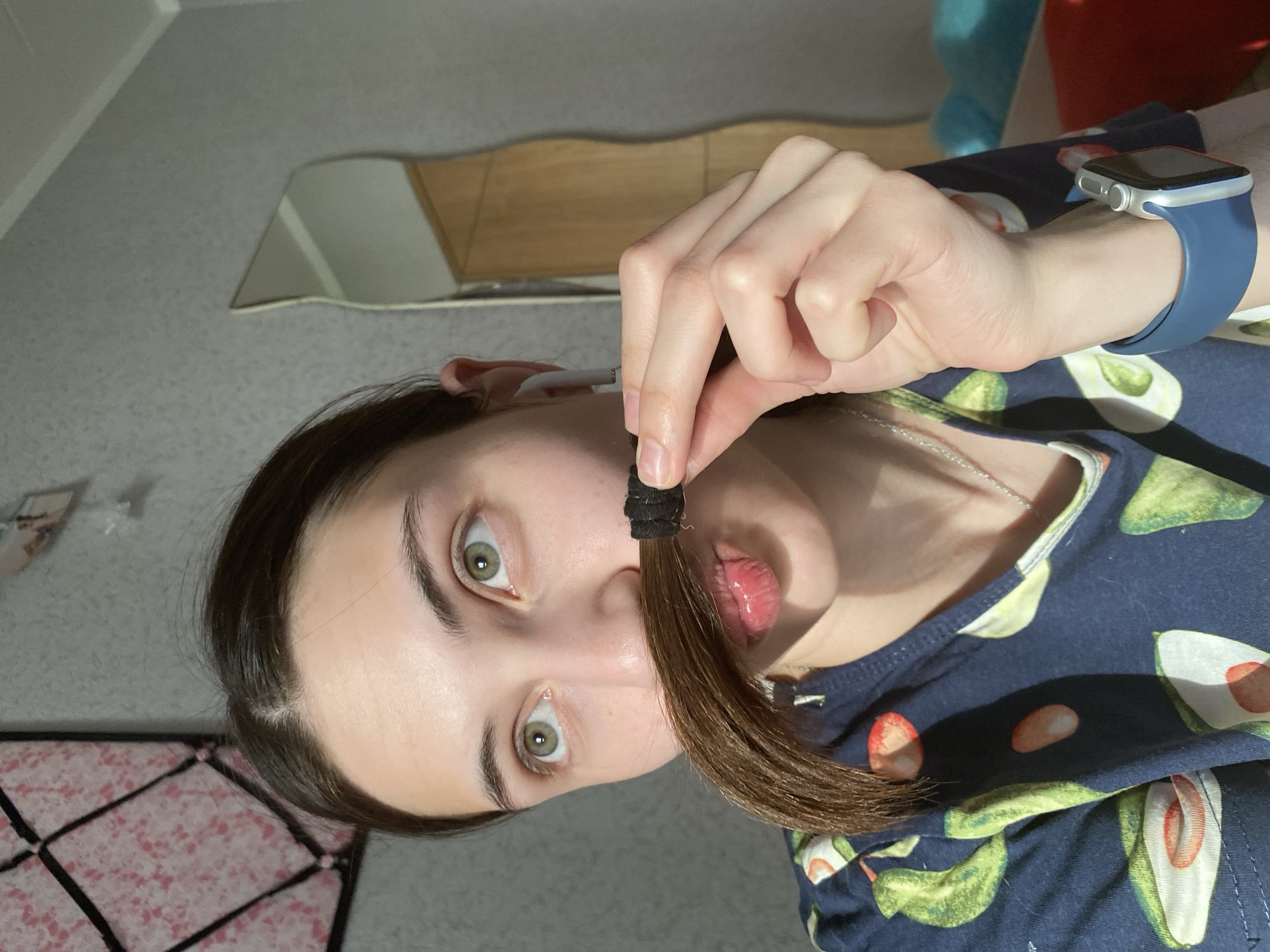A woman is holding a small ponytail of her brown hair with a black hair tie in front of her face. She is wearing a blue smartwatch with a blue band on her left wrist. She has light skin, green eyes, and dark hair. She is wearing a navy blue shirt with a pattern of avocados. In the background is a patterned gray and pink surface, likely a floor or rug, and further back is a partially visible wooden cabinet and a mirror on a gray wall. There is a partially visible teal object in the top right, and a possible white earbud in her left ear. There is nothing in the image to suggest a location in Votkinsk, Russia.