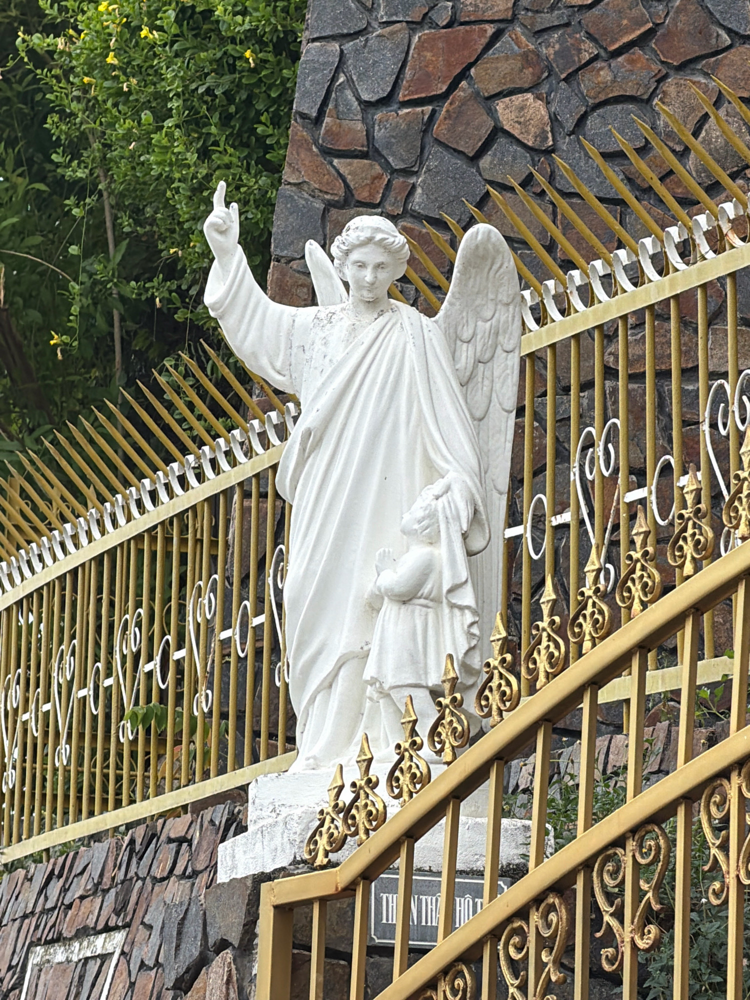 A white stone statue of an angel stands beside a gold metal fence. The angel is depicted with wings, a flowing robe, and is raising one hand with a pointed finger. Next to the angel's legs is a smaller white statue of a child. The fence has decorative scrollwork and pointed finials. Behind the fence and statue is a stone wall. Green foliage and yellow flowers are visible in the background. A sign with Vietnamese text is partially visible below the fence. The statue is located at the entrance to the Phat Trien Tinh Vien pagoda in Nha Trang, Vietnam.
