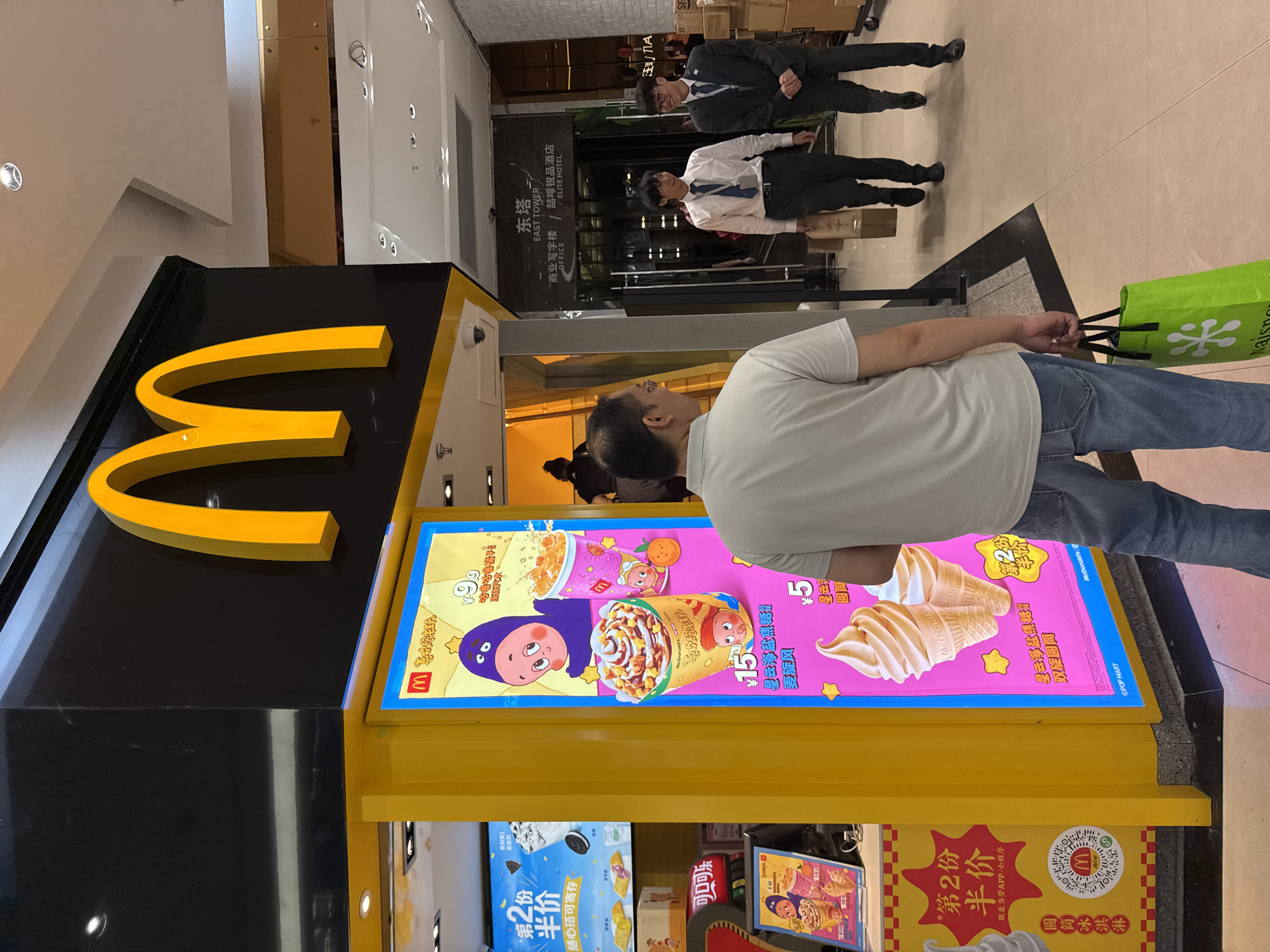 The image captures a McDonald's outlet, likely located within a commercial building in the Tianhe District of a Chinese city. The iconic golden arches of the McDonald's logo are prominently displayed. A large promotional display advertises ice cream products, featuring colorful cartoon characters and price points in Chinese Yuan. A man in a white shirt and jeans stands near the display, seemingly looking at it. Two men in business attire, one in a white shirt with a tie and the other in a suit, are walking in the background. The environment appears to be a well-lit indoor area, possibly a shopping mall or a commercial complex. In the distance, signage indicates the presence of an "EAST TOWER" and an "ELITE HOTEL." The floor is tiled, and the scene is generally clean and well-maintained.