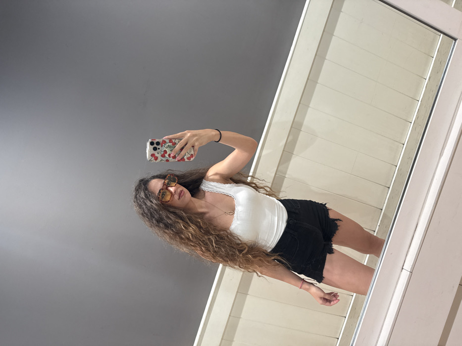 The scene is located in the city of Miami, United States of America. An individual is depicted taking a mirror selfie indoors. The individual is a female with long, wavy brown hair, wearing oversized tortoise-shell patterned sunglasses with yellow-tinted lenses. She wears a white ribbed tank top with lace trim along the neckline, black distressed denim shorts with frayed hems, and a gold-colored pendant necklace. A black hair tie is present on her left wrist, and a red bracelet on her right. She holds a silver-colored smartphone with a clear cherry-patterned case in her right hand, extended towards the mirror. The background consists of a plain gray wall above a light beige or off-white paneled wall, possibly wainscoting. A portion of the mirror's white frame is visible at the bottom.