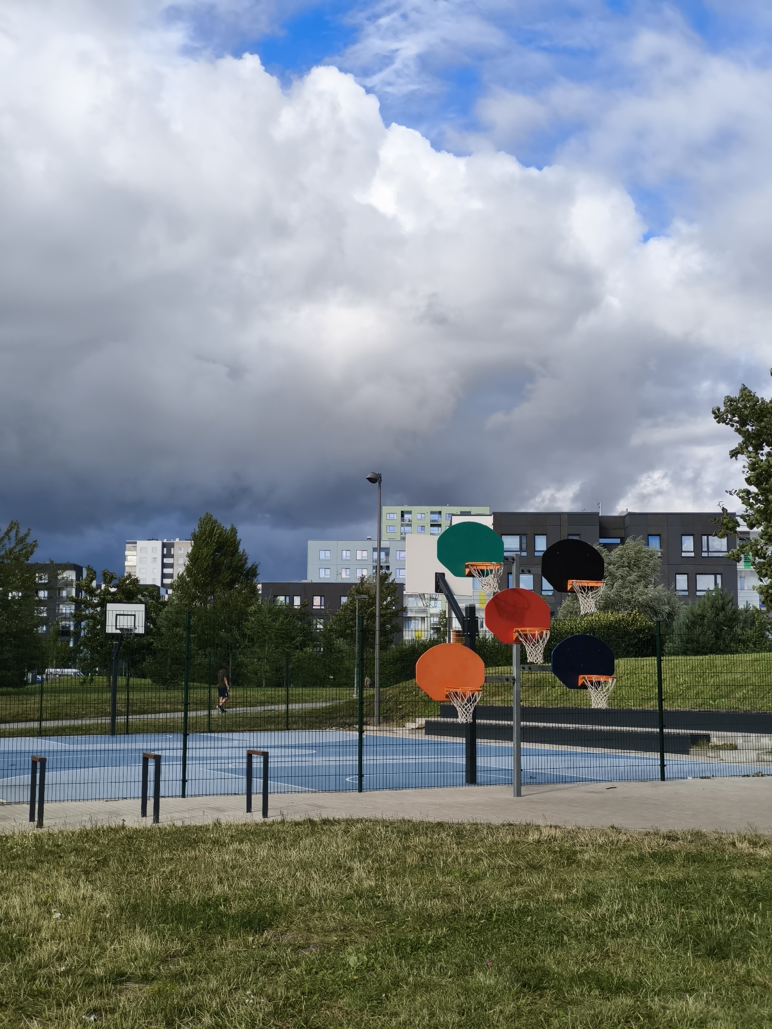 The image shows a basketball court in Tallinn, Estonia, with multiple colorful basketball hoops. In the background, there are modern residential buildings and lush green trees. The sky is partially cloudy, with a mix of white and dark gray clouds, suggesting an overcast day with the possibility of rain. A person is visible on the court, walking away from the camera. The court itself has a blue surface with white markings and is surrounded by a fence. The foreground consists of green grass. The overall scene depicts a recreational area within an urban environment.