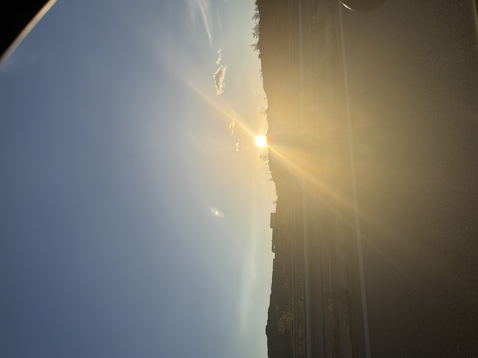 The image shows a bright sunburst over Belo Pole, Bulgaria. To the right, a structure casts long shadows, suggesting either buildings or infrastructure adjacent to the area where the sun sets. The sky is mostly clear, with a few wispy clouds near the sun.