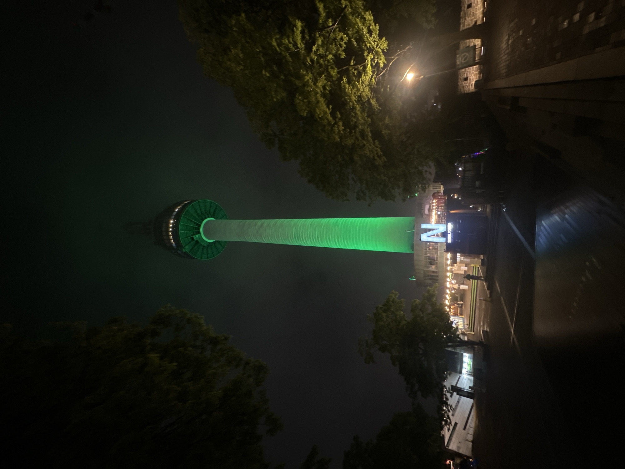 The image depicts the N Seoul Tower illuminated in green at night. The tower is centrally positioned and appears tall and cylindrical with a circular base. Trees frame the tower on both sides, and streetlights are visible in the distance. A building with illuminated signs is visible to the right of the tower's base, along with street-level activity involving pedestrians. The background is dark, suggesting a night scene.