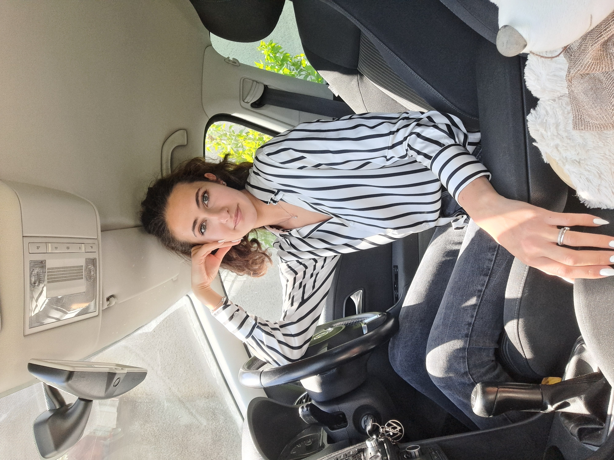 The image captures a young woman seated in the driver's seat of a car, looking directly at the camera. She has medium-length, curly brown hair and light-colored eyes, displaying a neutral yet engaging expression. She is wearing a black and white striped long-sleeved button-up shirt and dark jeans. A silver ring is visible on her left ring finger, and her fingernails are painted white. Her left hand is gently placed near her face, while her right hand rests on her lap.
The setting is the interior of a car, featuring a light-colored headliner and dark-colored seats and dashboard elements. Sunlight streams through the windows, indicating daytime, and green foliage can be seen outside, suggesting a pleasant outdoor environment. Visible car interior details include a rearview mirror, an overhead console with several buttons and lights (one of which is marked with a '0' and others with symbols), a black steering wheel, a gear shifter, and car keys hanging from the ignition. On the front passenger seat, a white, furry plush toy, possibly a bear, with a light brown nose and a small woven blanket or scarf is present. The overall atmosphere is calm and casual, with the woman posing for the shot.