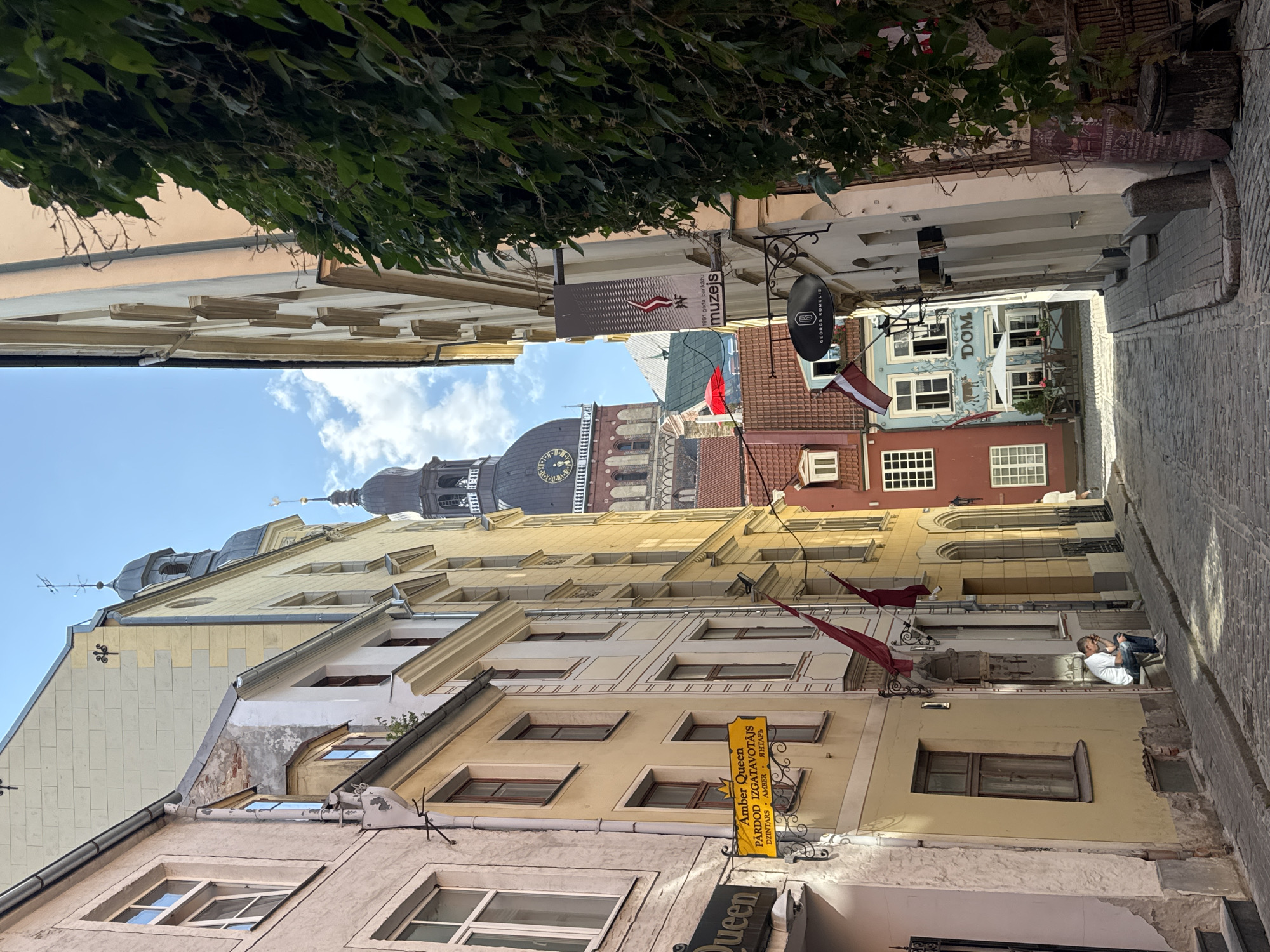 The image captures a narrow cobblestone street in Riga, Latvia, likely in the Old Town, as evidenced by the historic architecture and the prominent Riga Cathedral tower visible in the background. The street is lined with tall, narrow buildings of varying colors, predominantly yellow and cream, with some pastel pink and red facades. Green ivy cascades down the side of one building on the right.
Several signs are visible, including one for "Amber Queen" advertising "Dzintars Amber" and another for "muzejs" (museum). The Riga Cathedral tower, with its clock face, is a dominant feature in the distance, peaking above the buildings. Two Latvian flags hang from the facades of buildings.
In the foreground, on the left side of the street, a man is seated on a ledge, leaning against a yellow building, appearing to be contemplative or resting. He is wearing a white t-shirt and blue jeans. Further down the street, the facade of a building displays a sign with a cow illustration and the word "DOM" above it, suggesting a restaurant or shop. Another oval-shaped sign with the initials "G B" is mounted above it. The street is paved with cobblestones, indicating a historic setting. The sky is blue with scattered clouds, and the sunlight casts shadows, suggesting it is daytime.