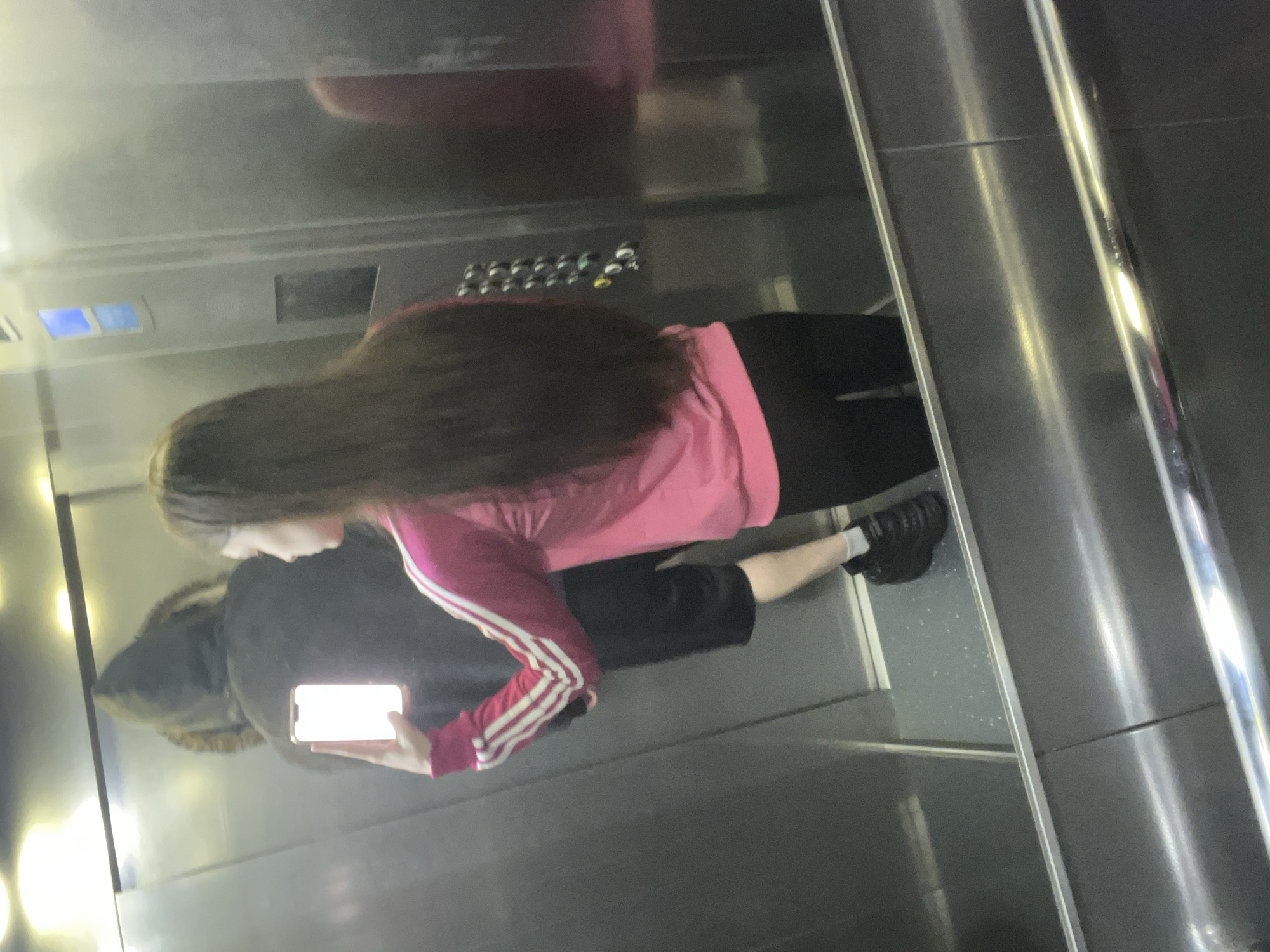 A person with long, dark hair, wearing a pink top with white stripes on the sleeves and black pants, is taking a picture of themself in a mirrored elevator. The elevator has control buttons and blue lighted indicators. The location and city are not ascertainable from the image.