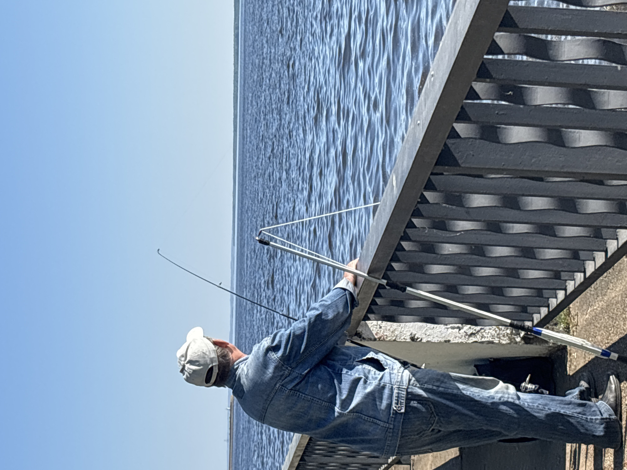 A man in a denim jacket and jeans, wearing a white cap, is fishing over a metal railing next to a body of water under a clear, blue sky. He holds a fishing rod in his hands. Another rod is propped against the railing. The scene is possibly in Chaikovsky, Russia, but no specific clues are present in the image to confirm this location.