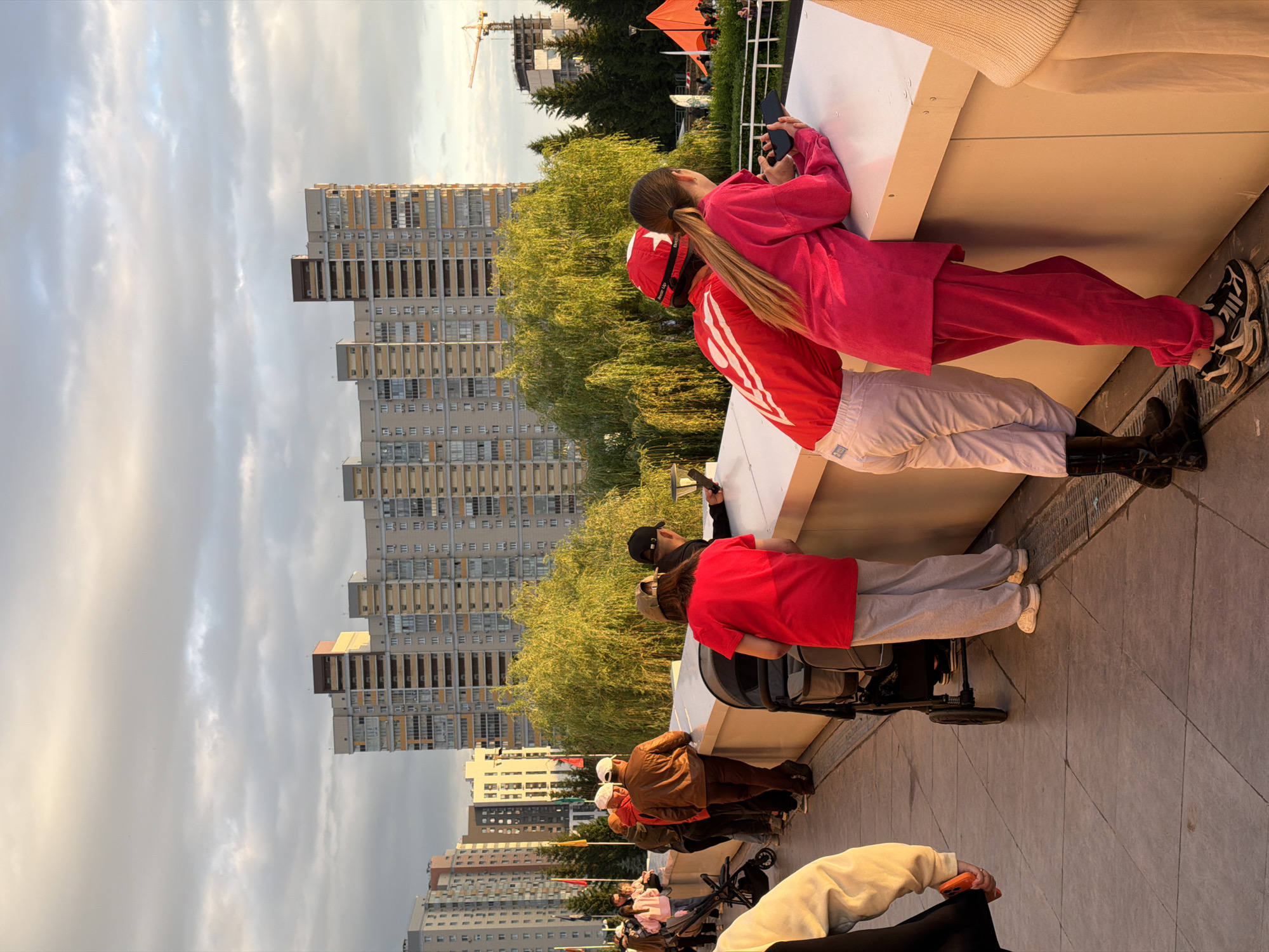 A group of people are standing on a paved walkway, looking over a white railing. In the background are several tall apartment buildings and trees. The sky is cloudy and the sun is setting, casting a warm light on the scene. One person in the foreground is wearing a red shirt and light-colored pants, and another is wearing a pink outfit. Another person in the middle is wearing a red helmet and a red and white shirt. There is a stroller on the left side of the image.
