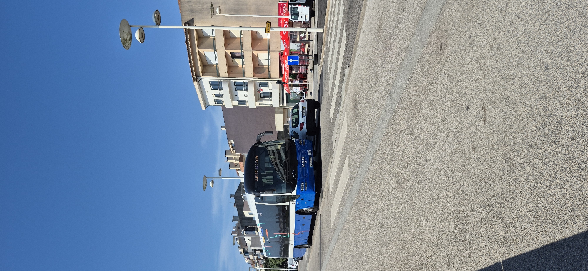 A blue and white MAN bus, with "unir" branding and the route number "3301 RU AGUÇADOURA" displayed, is stopped at a pedestrian crossing on a sunny day in Vila do Conde, Portugal. The bus is positioned on the right side of the road, facing forward. To the right of the bus, a white Peugeot 207 hatchback is also stopped at the crossing. In the background, there are multi-story buildings with balconies, and streetlights with distinctive circular shades are visible. A yellow sign with the word "Desvio" (Detour) and an arrow points to the right. A white van is parked further down the street. The road is paved and a pedestrian crossing is marked with white stripes. The sky is clear and blue. No people are clearly visible in action, though there might be people inside the shops or buildings. The scene depicts a typical urban street with public transportation and vehicular traffic.
