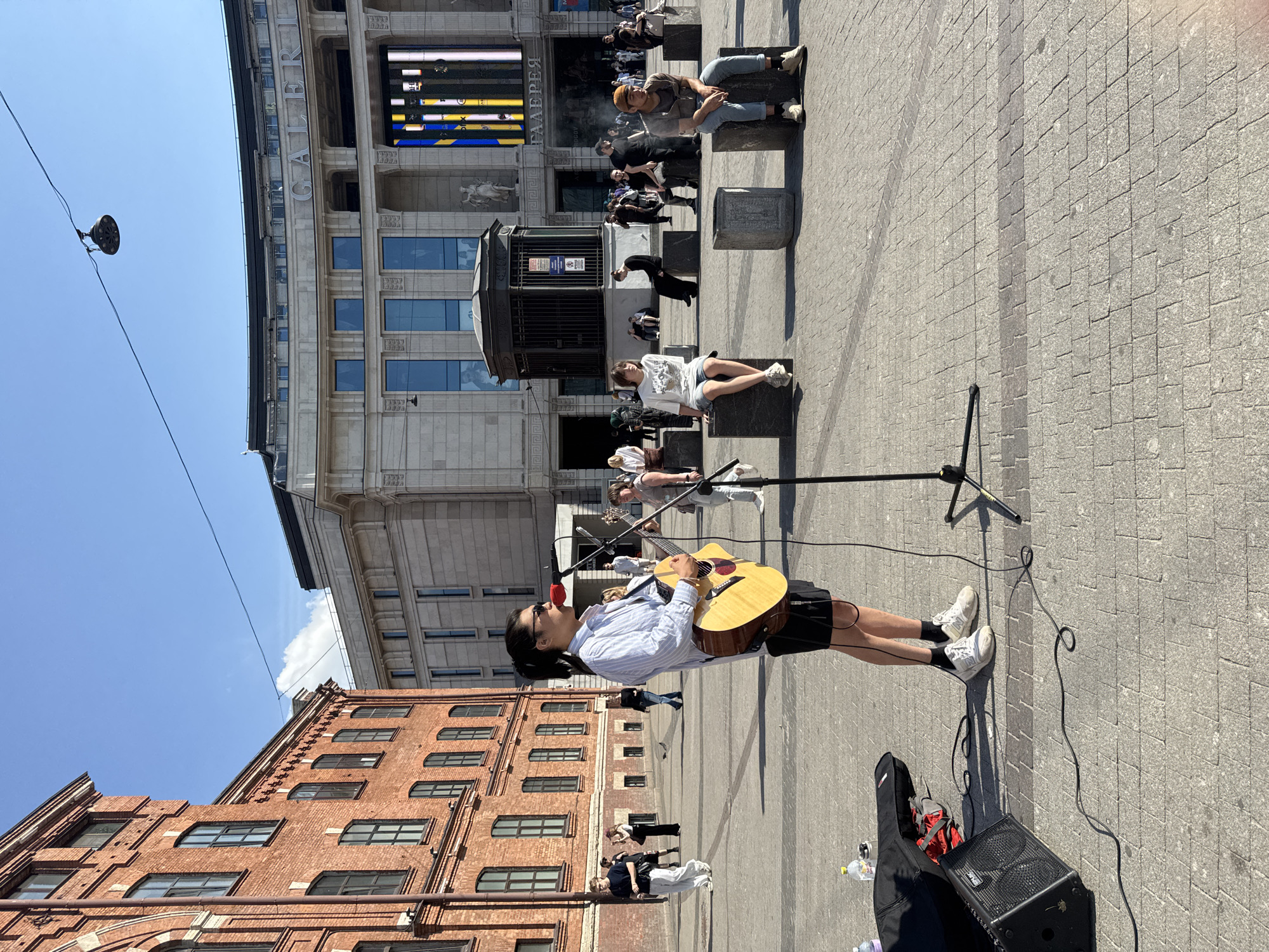 A street musician plays an acoustic guitar and sings into a microphone in a public square. The musician, dressed in a striped shirt, shorts, and sneakers, stands in the foreground. A guitar case and amplifier are on the ground beside him. In the background, other people are walking and sitting on cube-shaped benches. Behind them are buildings, including one with a prominent sign reading "GALEREYA," suggesting the location is in Saint Petersburg, Russia. The square is paved with interlocking stones. The sky is clear and blue, indicating a sunny day.
