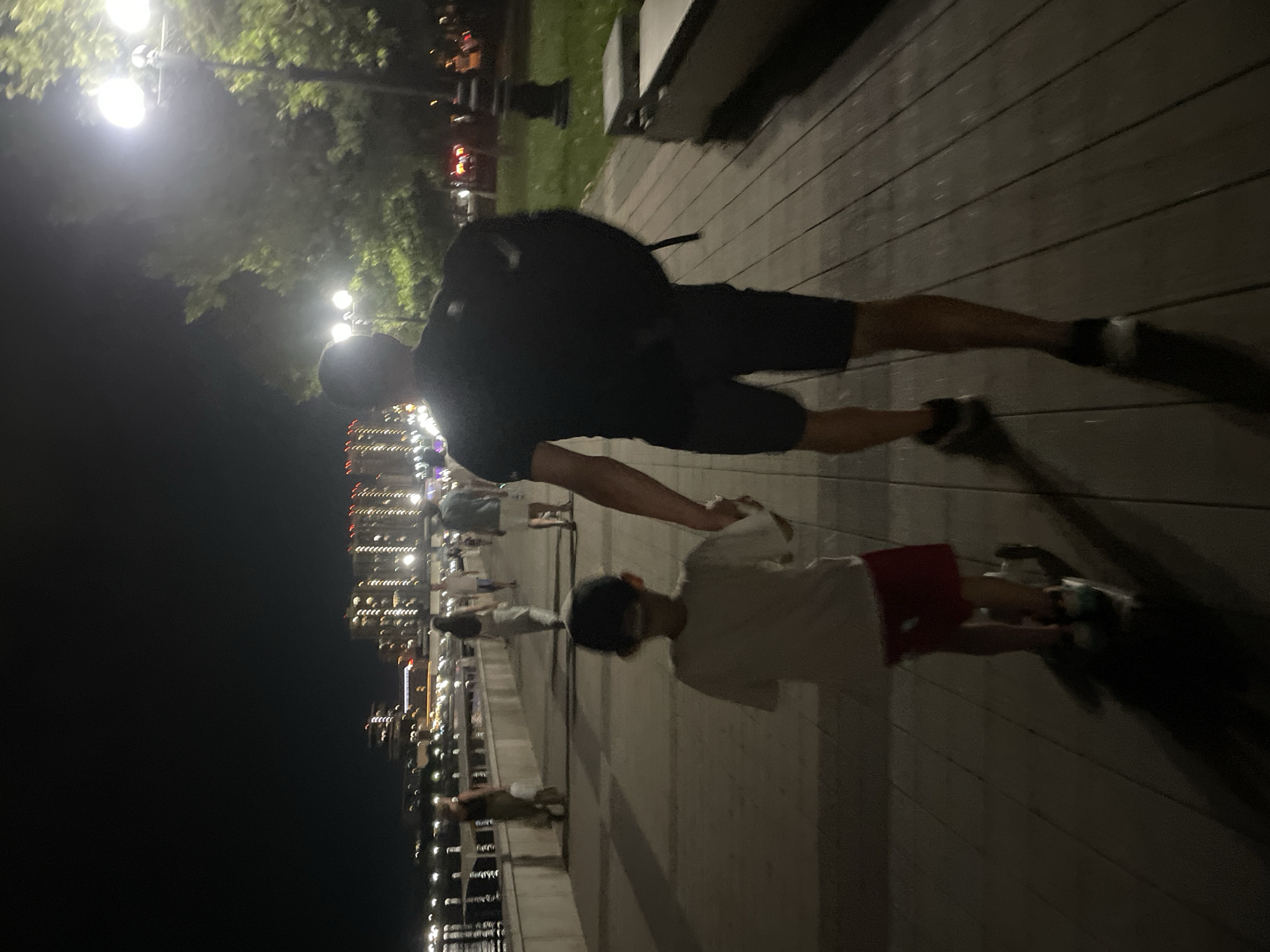 The image depicts an evening scene on a waterfront promenade in Sadovyi, Russia. A man, wearing a dark t-shirt and shorts, walks with a young boy. The boy, in a light-colored t-shirt and red shorts, is riding a scooter. The man's hand is clasped with the boy's. In the background, other people are visible strolling along the promenade. The promenade is paved with wooden planks and illuminated by streetlights and the lights from distant buildings across the water. The city skyline with its illuminated buildings is visible on the other side of the body of water. The overall atmosphere suggests a casual evening walk in an urban park setting.