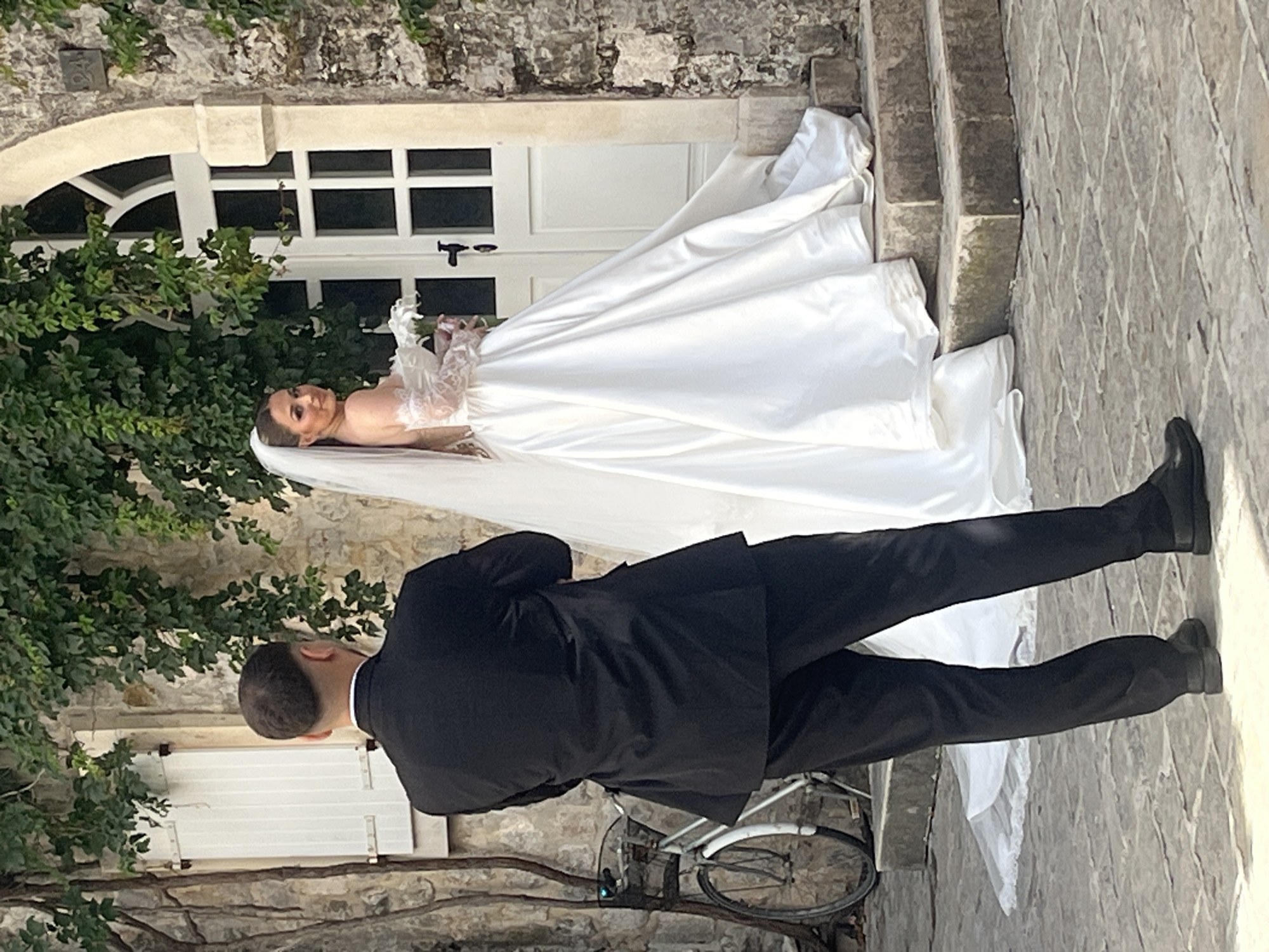 This outdoor photograph, set in Budva, Montenegro, captures a romantic wedding scene featuring a bride and groom. The image is vertically oriented, showing the groom in the foreground with his back to the camera, looking towards the bride who is positioned slightly elevated on a stone step.
The bride, the central focus, wears an elegant white wedding gown with a full, flowing satin skirt and a long train that cascades down the steps and onto the paved ground. Her bodice features intricate lace details and long, sheer lace sleeves. A white veil extends behind her, complementing her dark, styled hair. She looks towards the camera with a gentle smile, exuding happiness and serenity.
The groom is dressed in a classic black suit, white shirt, and black dress shoes. His posture suggests he is attentively observing or posing with the bride.
The setting is an old, charming European street or courtyard, typical of Budva's historic architecture. Behind the bride stands a light-colored stone building with a prominent white arched window and a white door with dark hardware. Lush green foliage, including ivy on the stone wall and a bush to the left, provides a natural, vibrant backdrop. The ground is paved with irregular stone tiles, adding to the rustic, historic atmosphere. A bicycle is partially visible in the lower background, leaning against the building.
The scene appears to be taking place during the daytime, under bright but soft natural light, suggesting pleasant weather suitable for an outdoor wedding photoshoot. There is no visible text in the image. The overall mood is celebratory and intimate, capturing a special moment between the couple.
