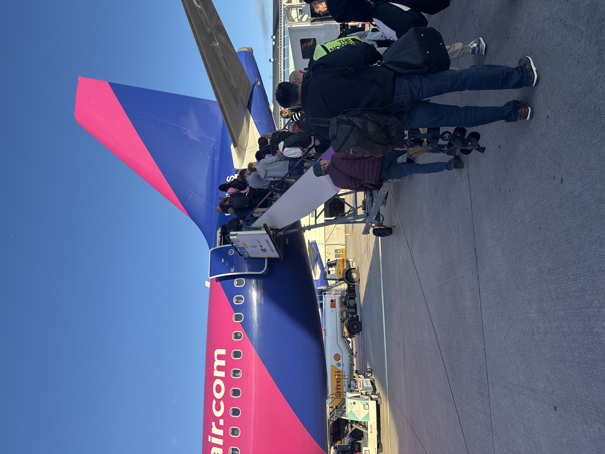 A group of passengers are boarding a Wizz Air airplane via a mobile stairway at an airport. The airplane is predominantly pink and blue, with "wizzair.com" branding visible on the fuselage. Several people are ascending the stairs, carrying luggage. On the tarmac, a fuel truck with "Shell" branding is parked near the plane. The sky is clear and blue.