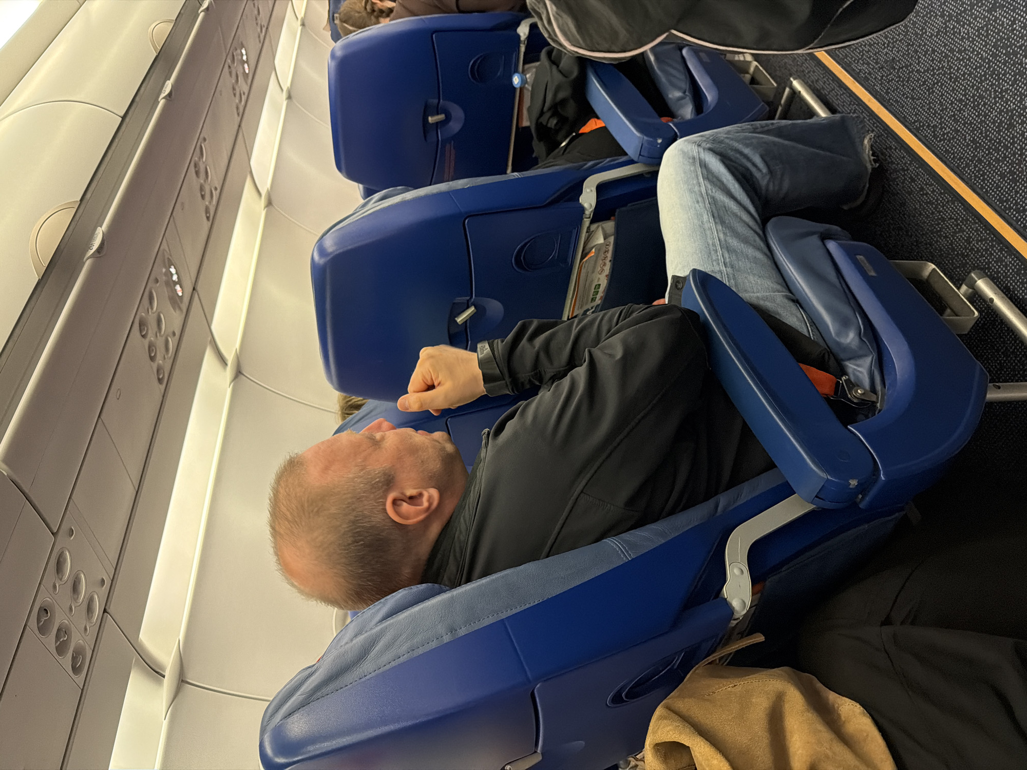 The image depicts the interior of an airplane cabin, with a focus on passengers seated in blue seats. In the foreground, a man with short, thinning hair is seated in a blue airplane seat, wearing a black jacket and blue jeans. He is looking to his right, with his right hand near his chin. To his left, a portion of a tan bag is visible.
Further back, more blue airplane seats are visible, some occupied and some empty. Luggage, including a black bag and a water bottle, is stowed in the seat pockets. The overhead compartment and lighting fixtures are also visible. The overall environment is that of a commercial airplane in flight. There is no visual information in the image that suggests the location is Saint Petersburg, Russia.