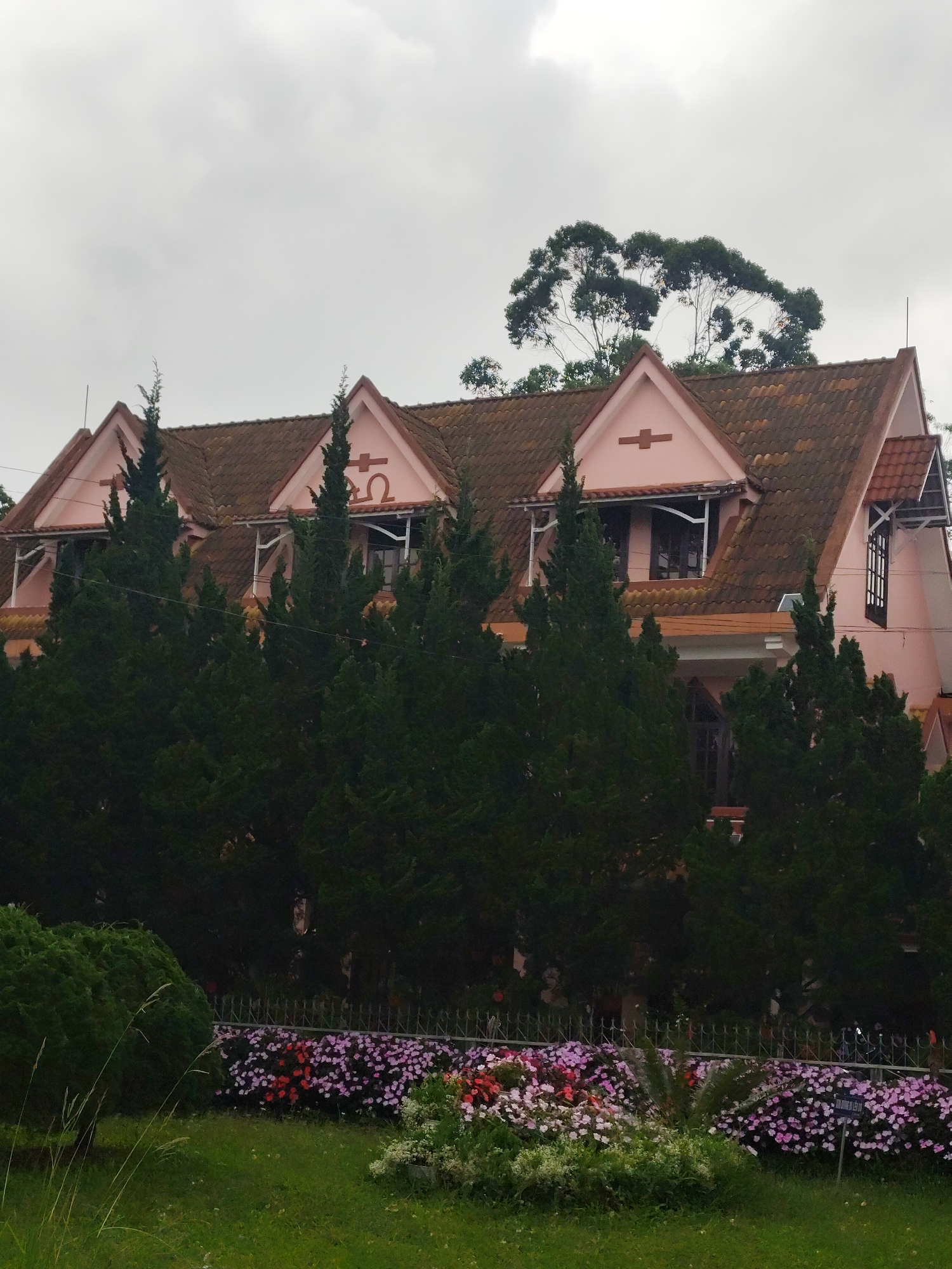 The image depicts a pink house with a terracotta tiled roof, featuring multiple gabled dormers with decorative symbols. The house is partially obscured by tall, dark green coniferous trees. In front of the trees, a low metal fence separates the house from a garden area. The garden contains a lush green lawn, a rounded green shrub, a dense flowerbed with a mix of pink, red, and white flowers, and a small palm-like plant. The sky is overcast and gray, suggesting a cloudy day. The scene is located in Cam Ly, Da Lat, Vietnam, a city known for its French colonial architecture and cool climate.
