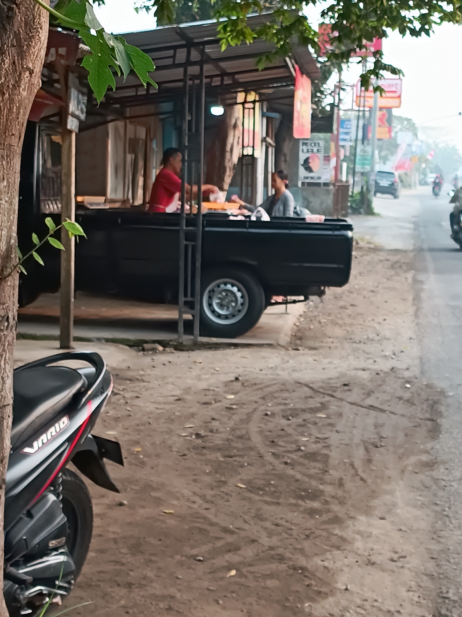 A low-angle, full shot captures a street scene in Jetis, Indonesia, focusing on a food stall set up beside a black pickup truck. A person in a red shirt is visible behind the truck, appearing to serve food to another person sitting on the truck's bed. The truck is parked on a dirt road, with a motorcycle in the foreground. The background shows a street with more stalls and vehicles, suggesting a busy market or roadside eatery. The lighting suggests it is daytime, possibly late morning or early afternoon.