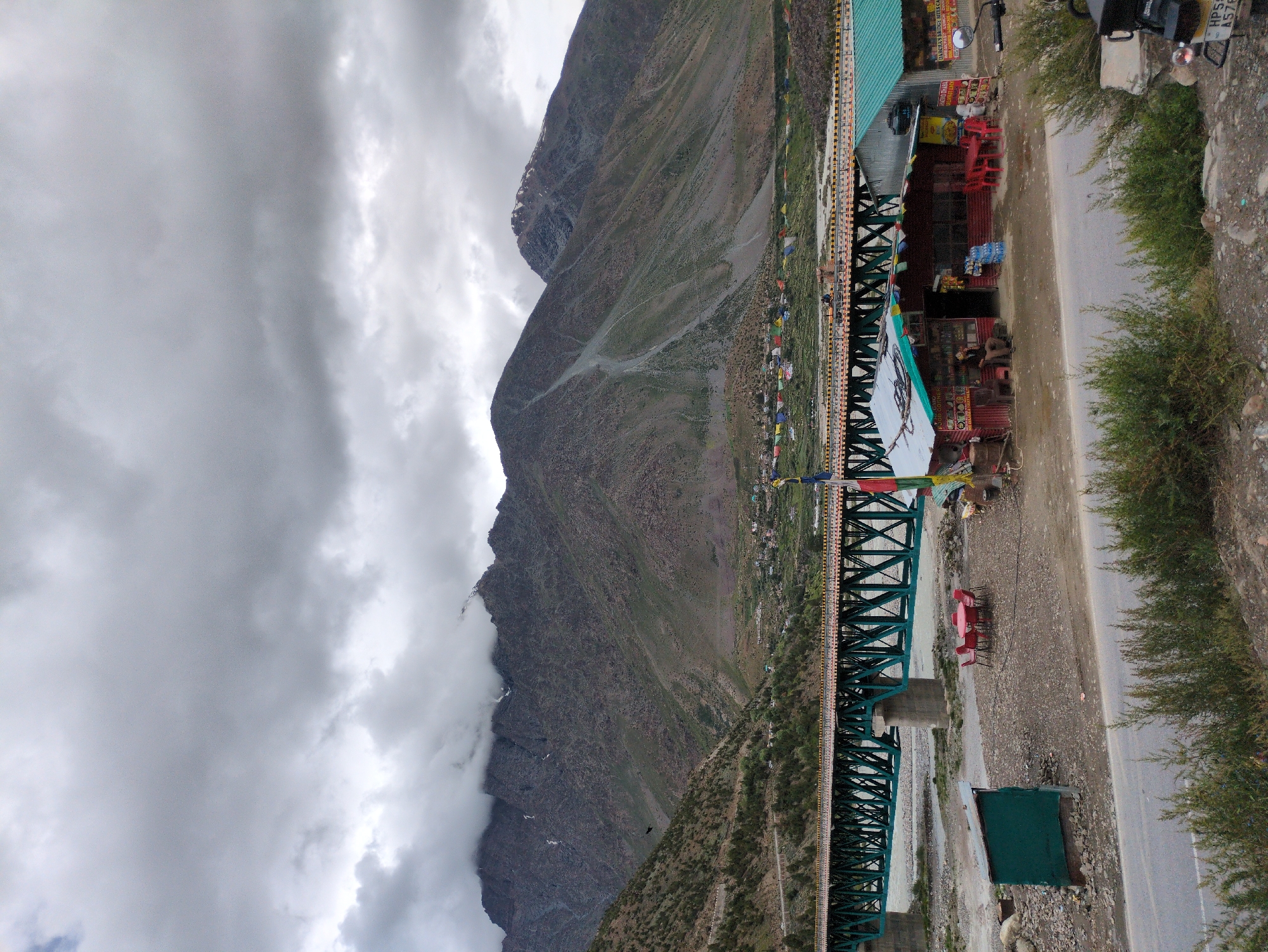 The image depicts a roadside scene in Darcha, India, with a focus on a small cluster of roadside eateries and shops. A long, green truss bridge spans a river in the background, with steep, rocky mountains rising behind it. The sky is overcast with heavy clouds, suggesting a potential for rain or a generally gloomy atmosphere.
In the foreground, a paved road runs horizontally across the bottom of the image. A motorcycle is parked on the right side, partially visible. To the left of the road, several small shops and food stalls are lined up. One prominent red building with a green roof has signs indicating it is a food corner. A person, possibly an employee or customer, is seated on a chair outside this establishment. Red tables and chairs are set up outside some of the shops, suggesting outdoor seating for customers. A small, green shed is also present to the left.
Further back, the truss bridge is a dominant feature, its green structure contrasting with the muted tones of the landscape. The river flows beneath it, its banks appearing rocky and sparsely vegetated. On the opposite side of the river, a small village or settlement is visible nestled at the base of the mountains, with colorful prayer flags strung across the landscape. The mountains themselves are rugged and imposing, with patches of vegetation and scree slopes. Some snow can be seen on the higher peaks. The overall impression is one of a remote, mountainous region with essential services catering to travelers.