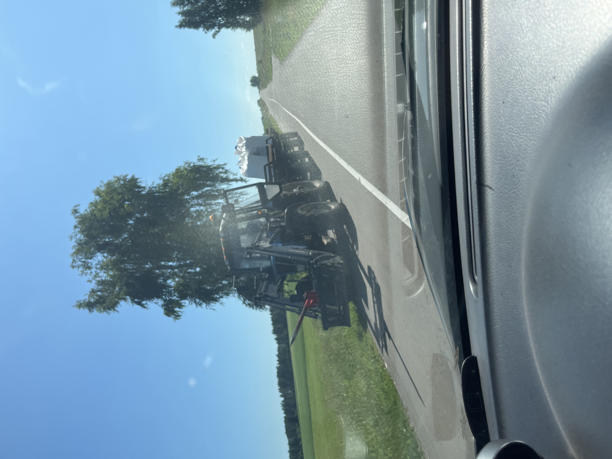 The image shows a blue tractor with a trailer carrying a large white object, possibly a bale of hay or a large package, driving on a paved road. The tractor is on the right side of the road, moving away from the viewer. The road is bordered by a green grassy field on the left and more grass and trees on the right. The sky is clear and blue. The image appears to be taken from inside a vehicle, with the dashboard visible in the foreground. The location is identified as Tuksy, Russia.