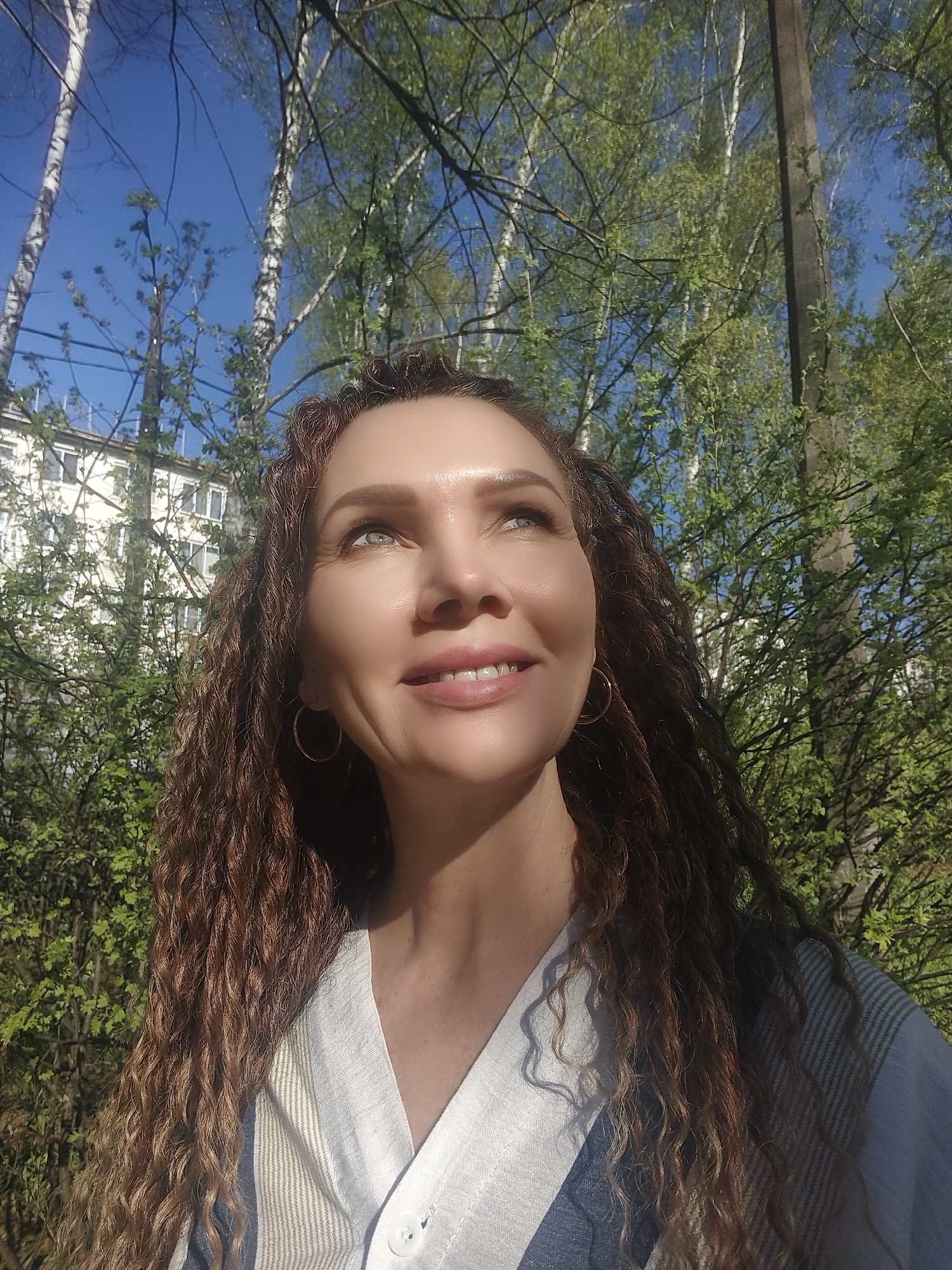 A medium shot depicts a woman outdoors. She has fair skin, light eyes, and long, curly brown hair. She is smiling and looking upwards, with light reflected on her face. She wears gold hoop earrings and a white shirt with a button closure.
Behind her, a building is partially visible to the left, surrounded by trees with green leaves. Several birch trees are also visible, with their distinctive white bark. The sky is blue, indicating a clear day. The scene appears to be a park or garden setting in an urban area.