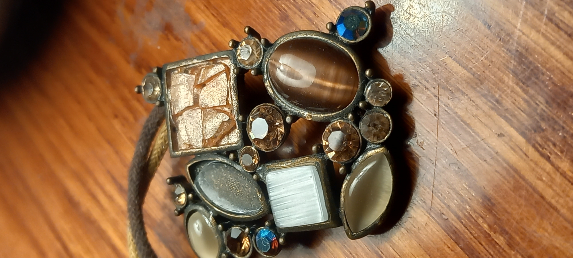 The image presents a close-up, elevated view of an ornate, vintage-style piece of jewelry resting on a warm-toned wooden surface. The primary subject is a multi-gemstone embellishment set in an antique bronze or brass-colored metal.
The jewelry piece is composed of various shapes and colors of stones. A prominent oval, brown-gold stone with a chatoyant cat's eye effect is centrally positioned, alongside a large square stone exhibiting a textured, cracked, or gold-leaf-like golden-brown appearance. Surrounding these are numerous smaller, faceted stones, predominantly round, in shades of brown, amber, and clear. Two small, vibrant blue round stones add contrasting color, one at the top right and another towards the bottom left. Other notable elements include a rectangular white stone with horizontal striations, possibly mother-of-pearl or shell, and several marquise-shaped and oval stones in light grey and beige tones, some appearing translucent. A darker grey marquise-shaped stone also forms part of the design. A braided cord or strap, in shades of brown and gold, is partially visible on the left side, suggesting the item may be part of a necklace, bracelet, or an adornment for another accessory.
The background consists of a wooden surface, possibly a tabletop or floorboard, characterized by a visible grain pattern and numerous scratches and signs of wear, indicating age or frequent use. The lighting casts subtle shadows, enhancing the depth and texture of the jewelry. No specific activity or event is taking place, as the object is stationary. There is no visible text within the image. The location context provided for this media file is Kryvyi Rih, Ukraine.