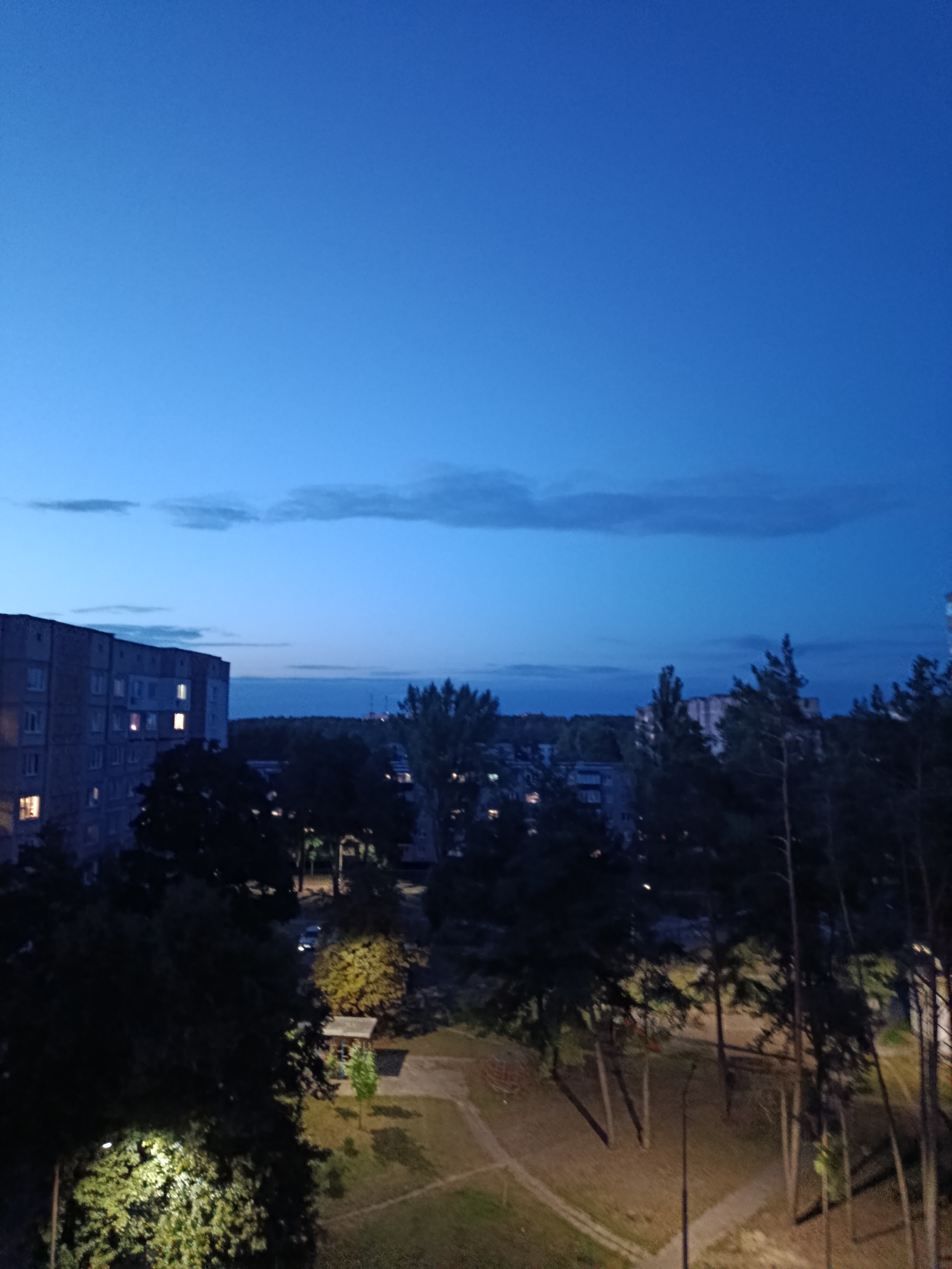 The image depicts a nighttime view over a residential area in Svietlahorsk, Belarus. In the foreground, to the left, a multi-story building with lit windows is visible, suggesting an apartment complex. Dark trees dominate the middle ground, with some of their branches casting shadows. A paved pathway and a small playground area are discernible on the ground, illuminated by streetlights. In the distance, more buildings and trees can be seen, along with a sliver of the horizon. The sky is a deep blue, transitioning to a lighter shade near the horizon, with faint clouds. A single car is visible on a road in the middle ground, its headlights on.