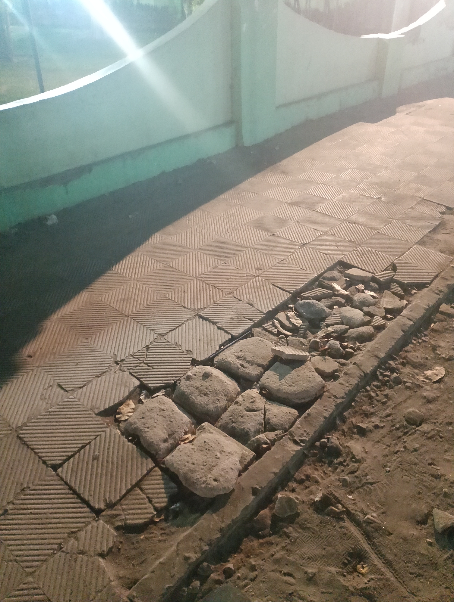An outdoor shot captures a damaged section of a tiled walkway, likely in Ngadiluwih, Indonesia. The walkway is composed of square tiles with a ribbed pattern, arranged in a diamond formation. A portion of the walkway has collapsed, revealing a gap filled with rocks and dirt. The surrounding area appears to be a dirt path, with some scattered leaves. In the background, a light green wall with a curved top is visible, and a bright light source casts strong diagonal shadows across the scene. The overall impression is one of disrepair and neglect.
