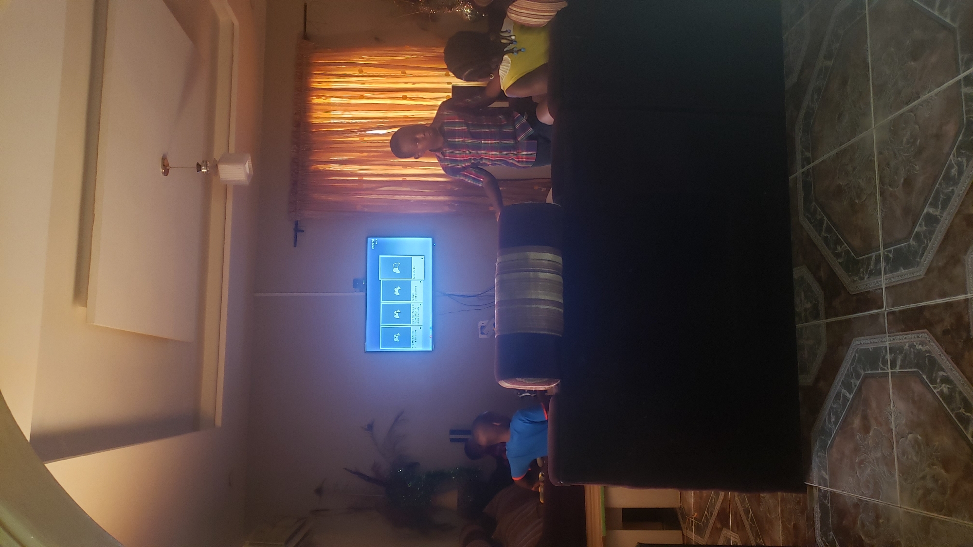 The image shows an interior scene in Sabon Tasha, Nigeria. There are three children present, positioned on and near a dark-colored sofa. One child, wearing a red and blue plaid shirt, stands near a television that displays what appears to be a gaming menu. A second child, in a green shirt, is partially visible behind the sofa. A third child is seen wearing a blue shirt. The room features decorative ceiling elements and a window covered with orange curtains. The floor is tiled with a pattern of octagons and squares. A cylindrical object is situated on the sofa. A houseplant is also visible in the room.