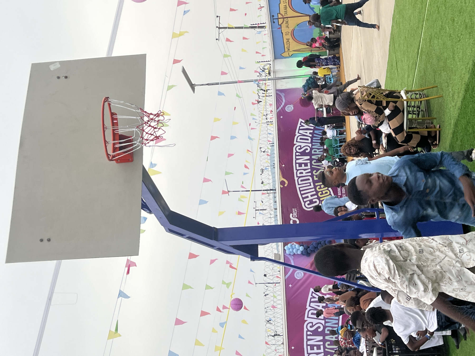 The image depicts an outdoor event likely related to Children's Day. In the foreground is a basketball hoop with a white backboard, a red ring, and a white and red net. The hoop is mounted on a blue support structure. A line of colorful pennant flags is strung across the background. A large banner with "CHILDREN'S DAY...S/CARNIVAL" is partially visible. Beyond the banner, a crowd of people is visible. Some are seated on chairs, while others are standing. The background also shows a building or structure with "WELCOME TO JALINGO TARABA STA" written on it. A pink ball is suspended in the air near the basketball hoop. The city and country are not visible in the image, so these can not be described.